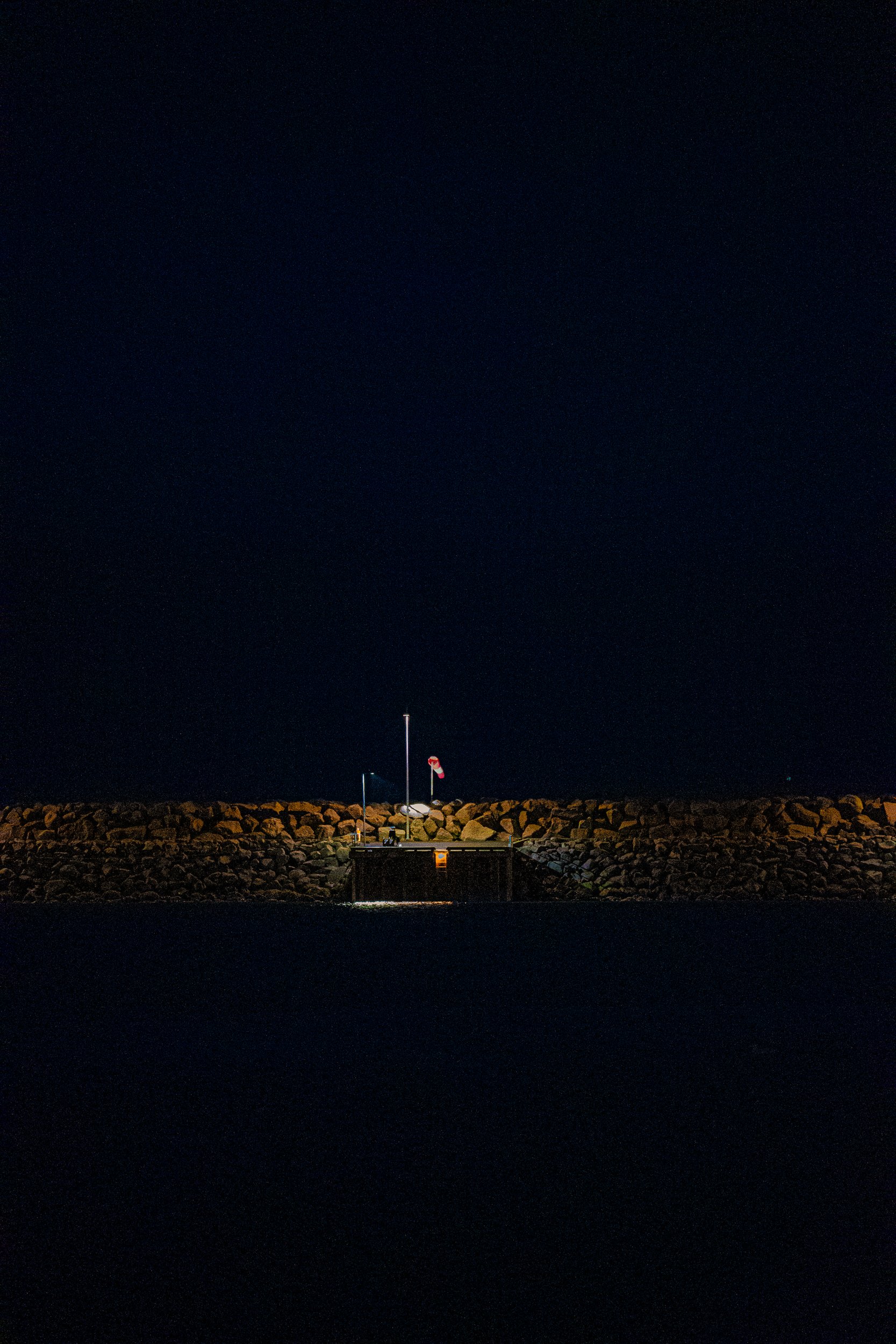 Nighttime view of a rocky breakwater with a lit navigation platform, flagpole, and windsock over dark water