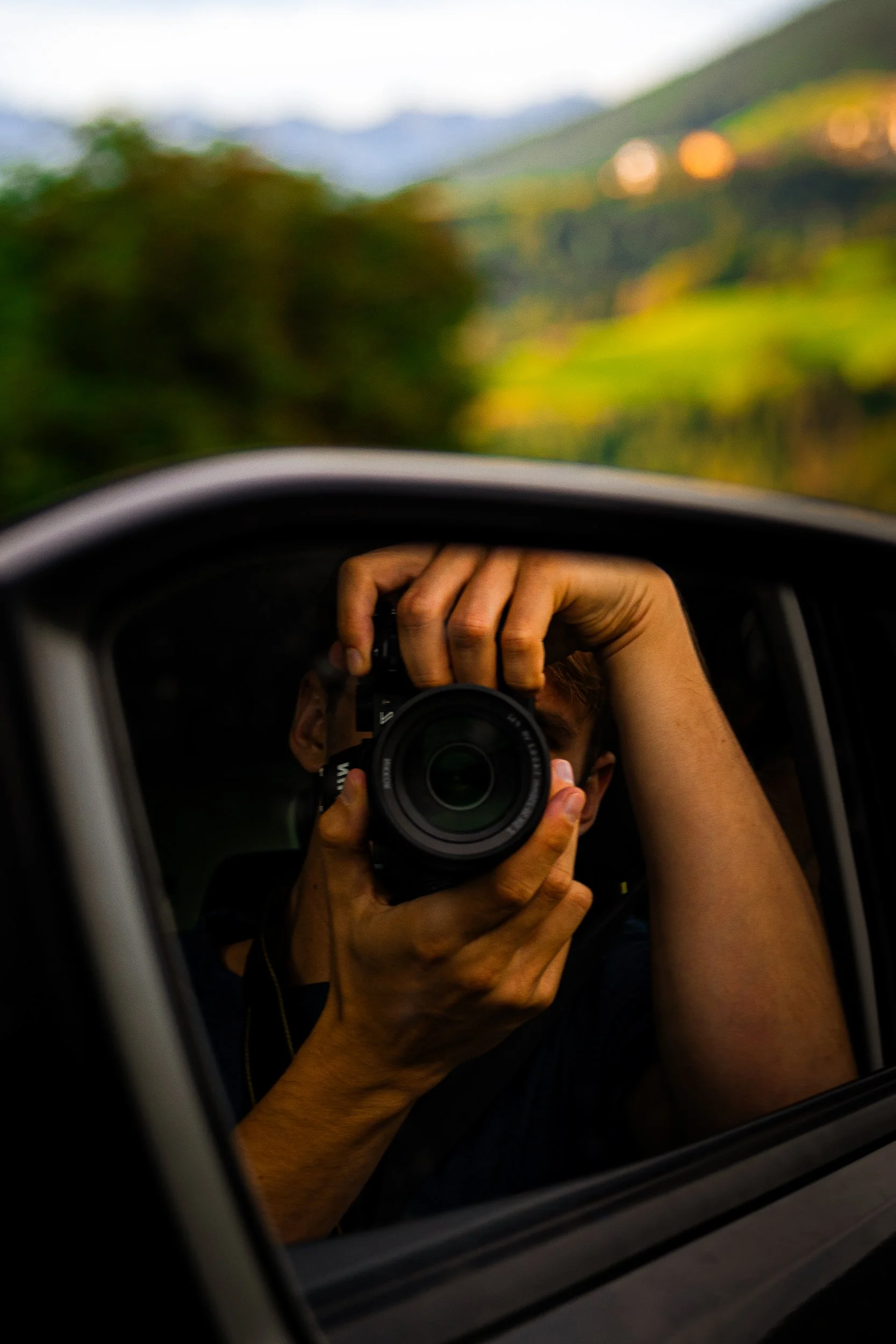 Photographer holding a camera while taking a self-portrait through a car window with a blurred mountain landscape outside