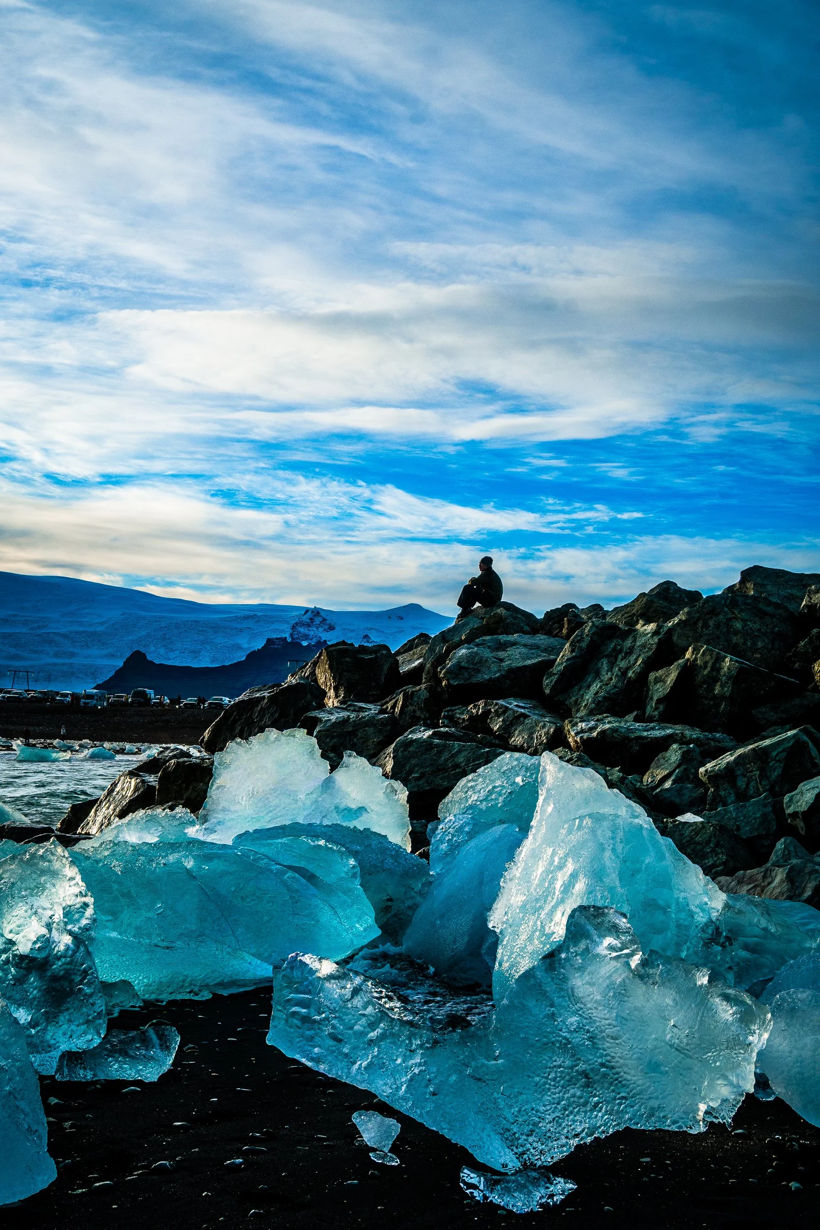 Tourist sitting on volcanic rocks above blue ice chunks on Iceland's black sand Diamond Beach