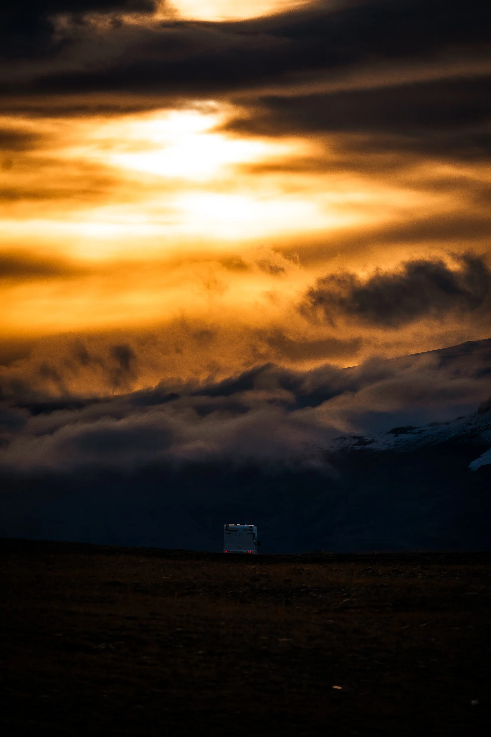 White camper van silhouetted against glowing orange storm clouds over a dark mountain landscape