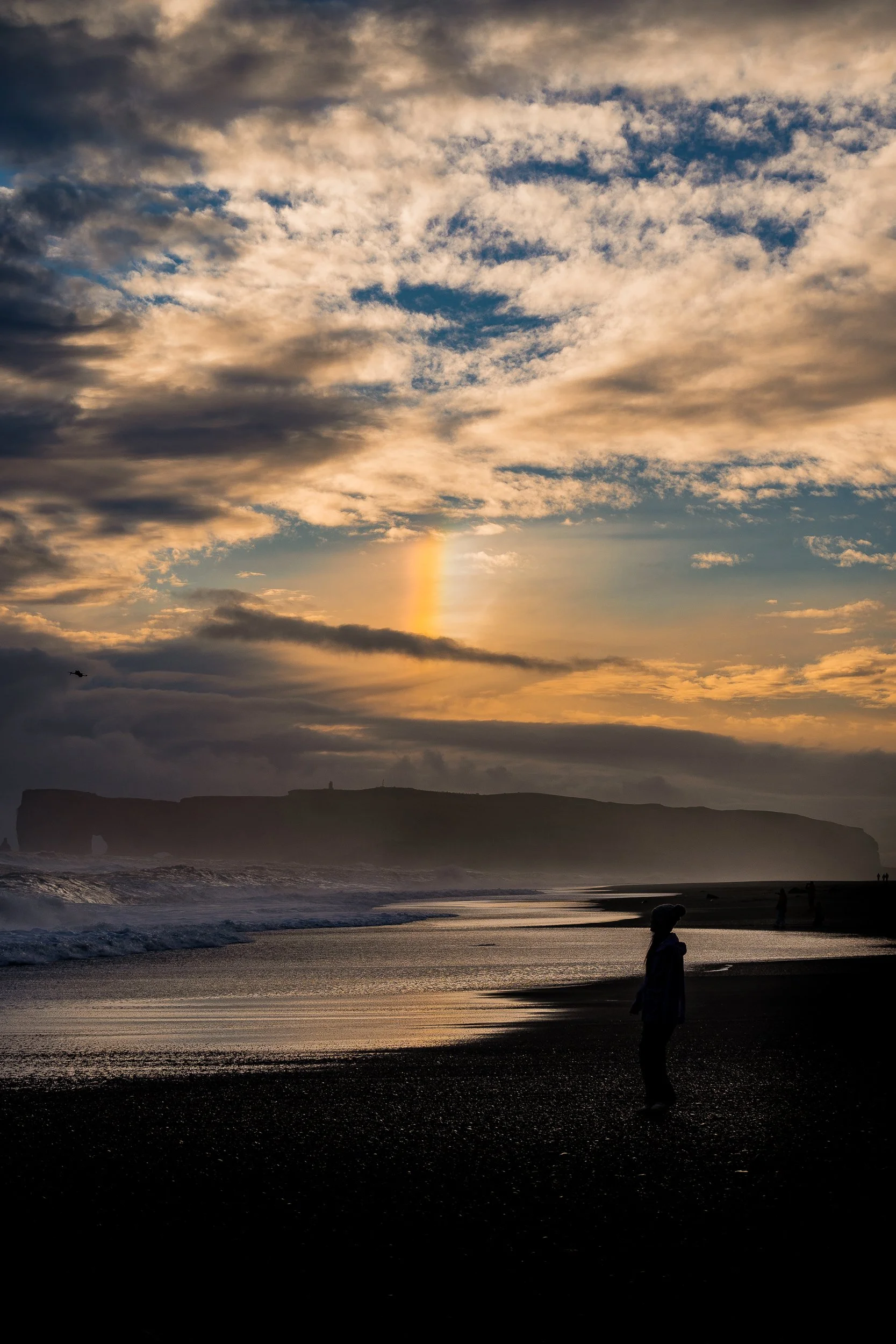 Silhouetted person on a black sand beach at sunset, with ocean waves, cliffs, dramatic clouds, and a distant bird.