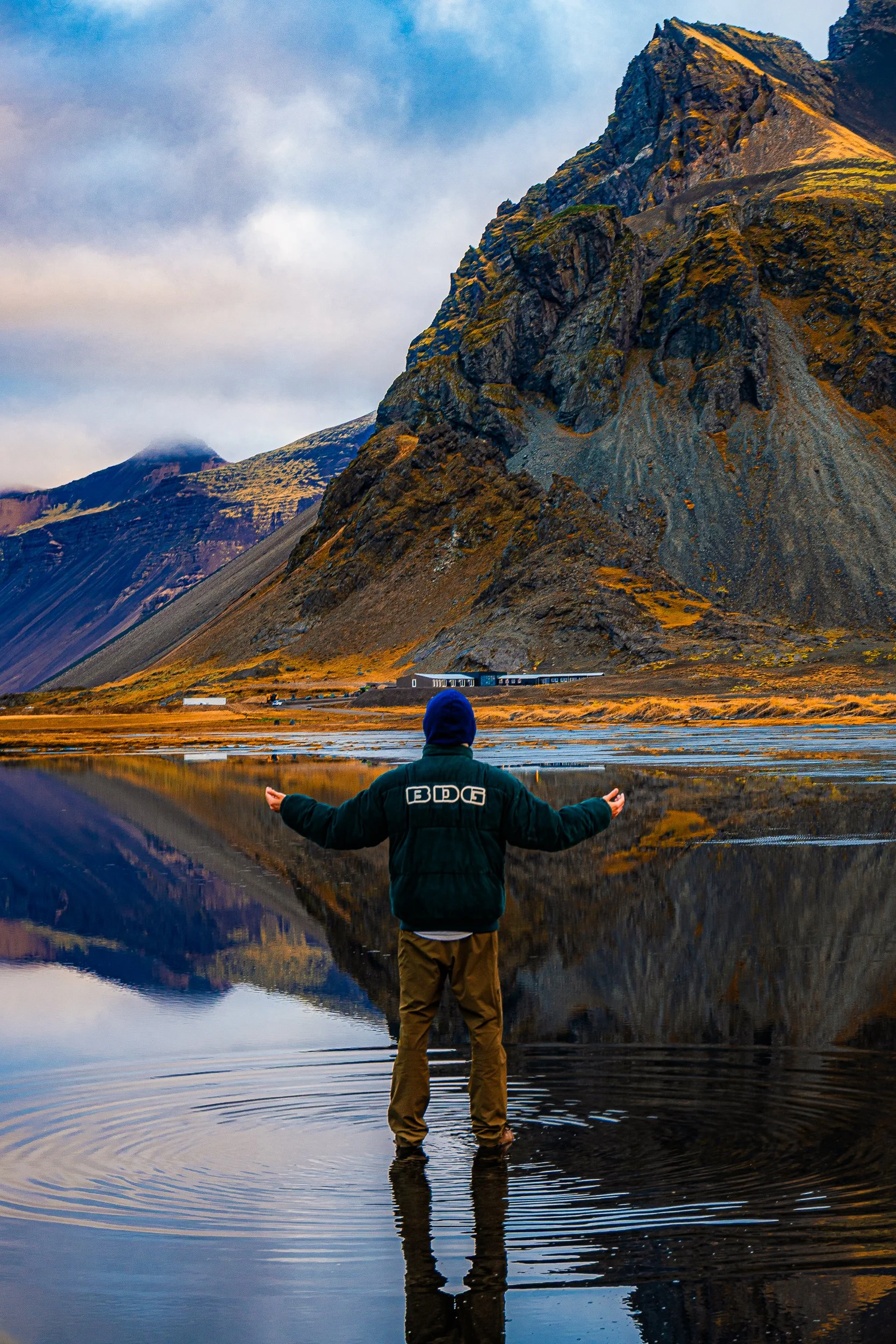 Person standing in a mountain lake with arms outstretched, facing rugged peaks and their reflections.