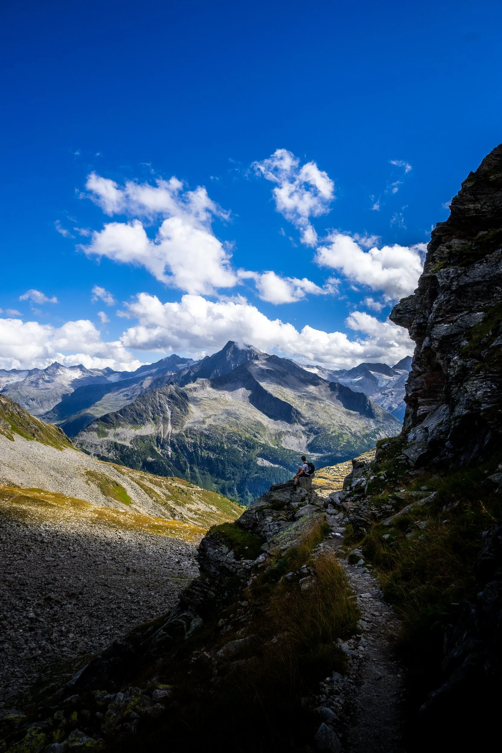 Hiker resting on a rocky mountain trail overlooking alpine peaks beneath a bright blue, cloud-filled sky