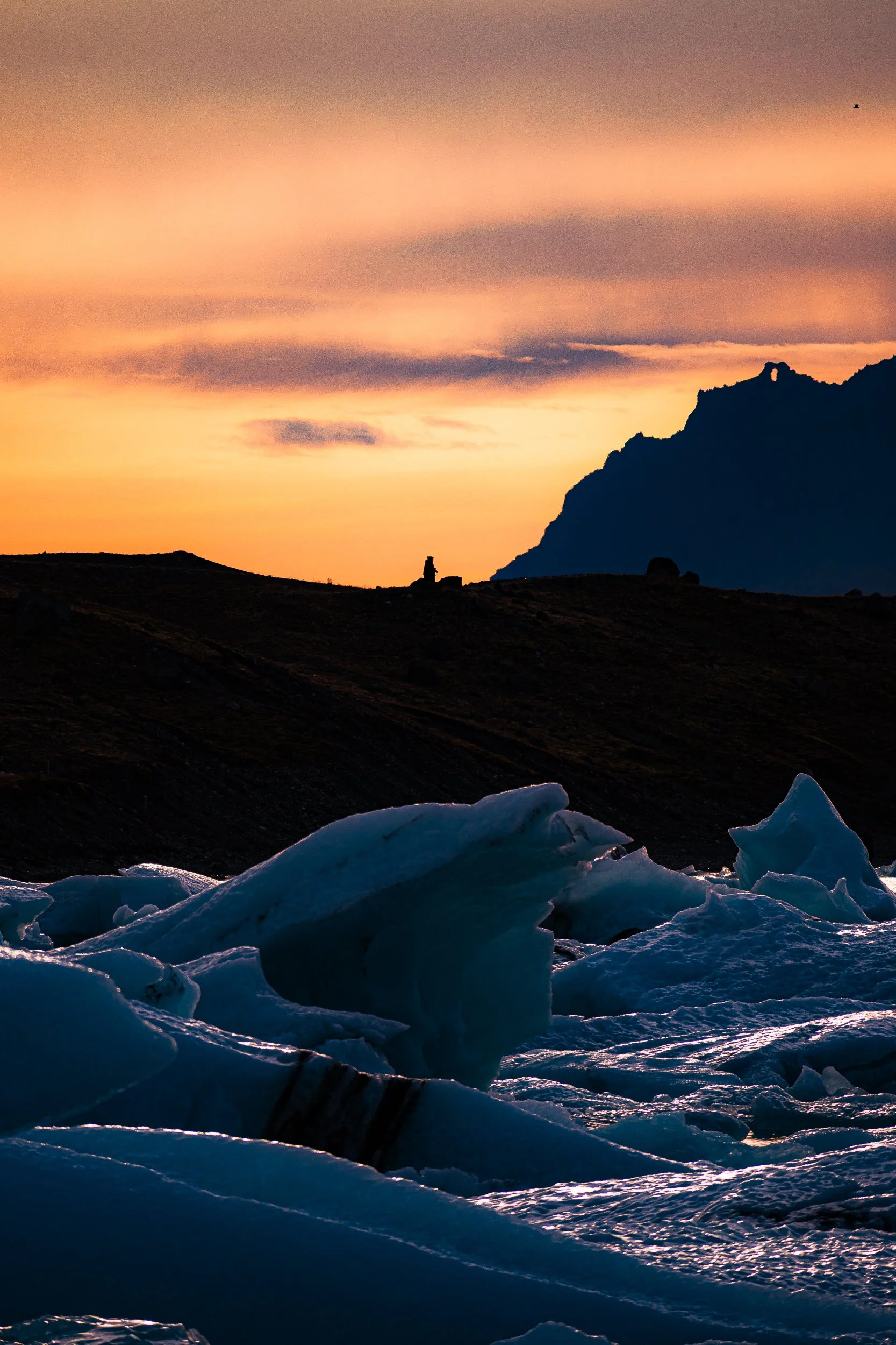 Jagged blue ice floes in the foreground beneath a silhouetted mountain and glowing orange sunset sky