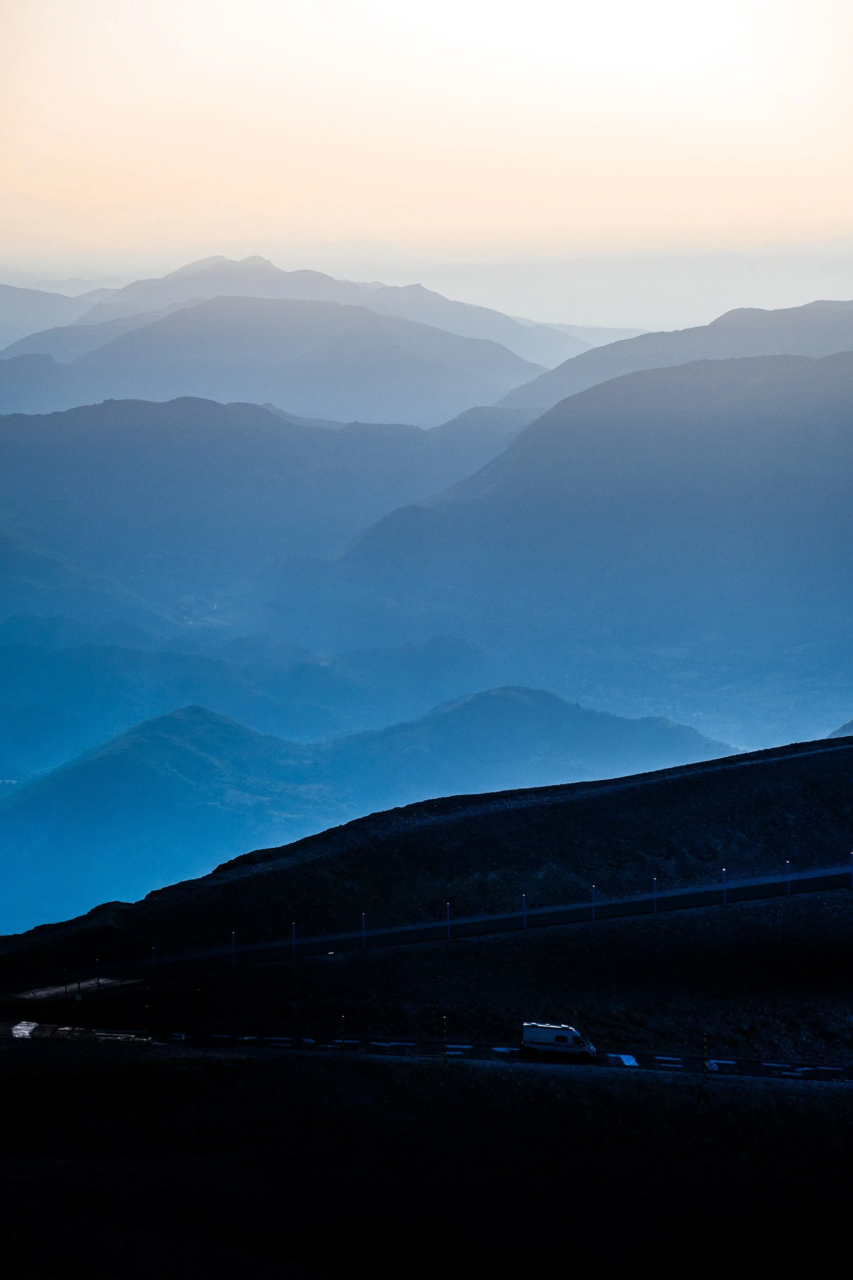 Layered blue mountain ranges at dawn with a winding mountain road and van in the foreground