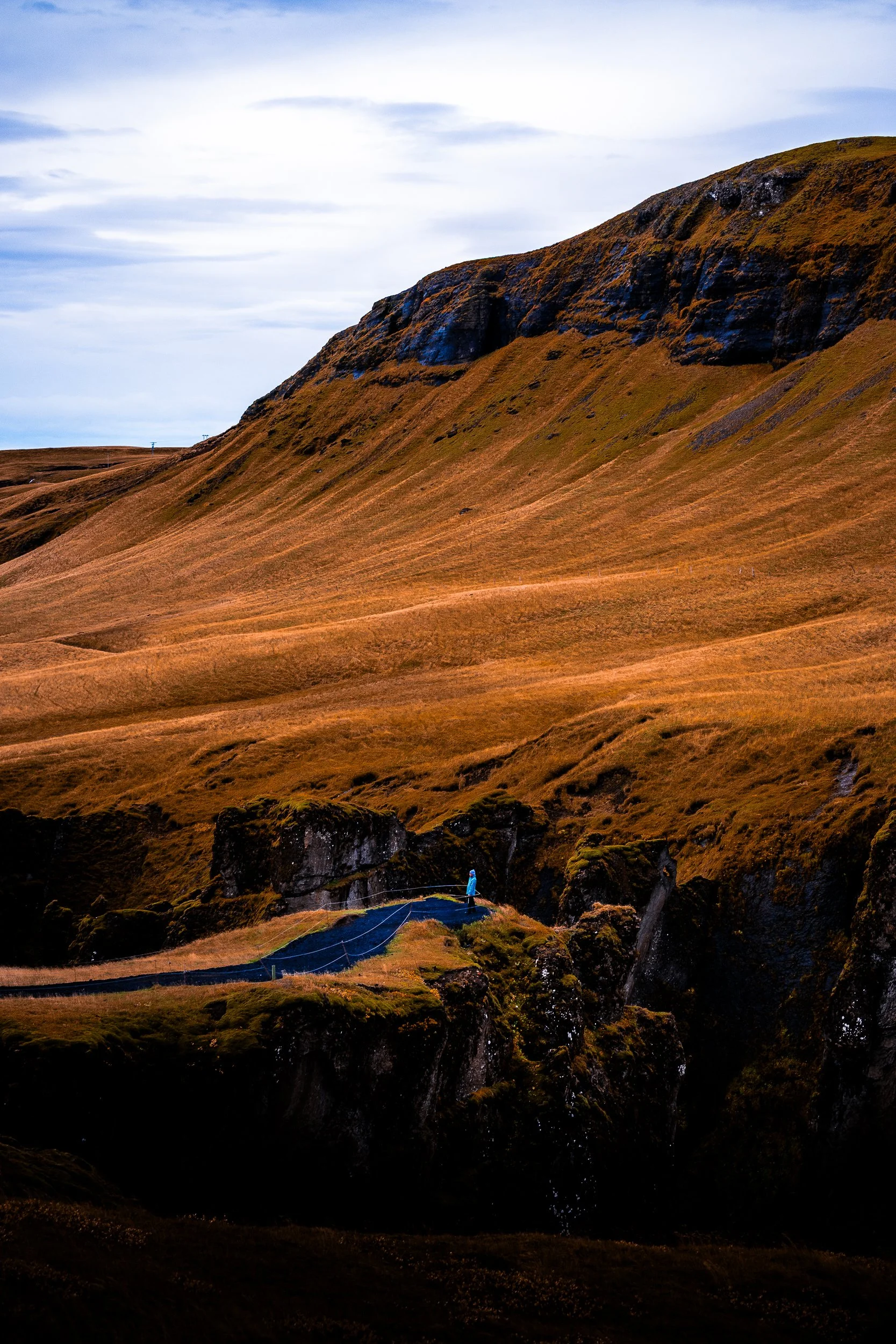 Hiker on a fenced trail through moss-covered volcanic cliffs and golden grassland beneath a cloudy sky