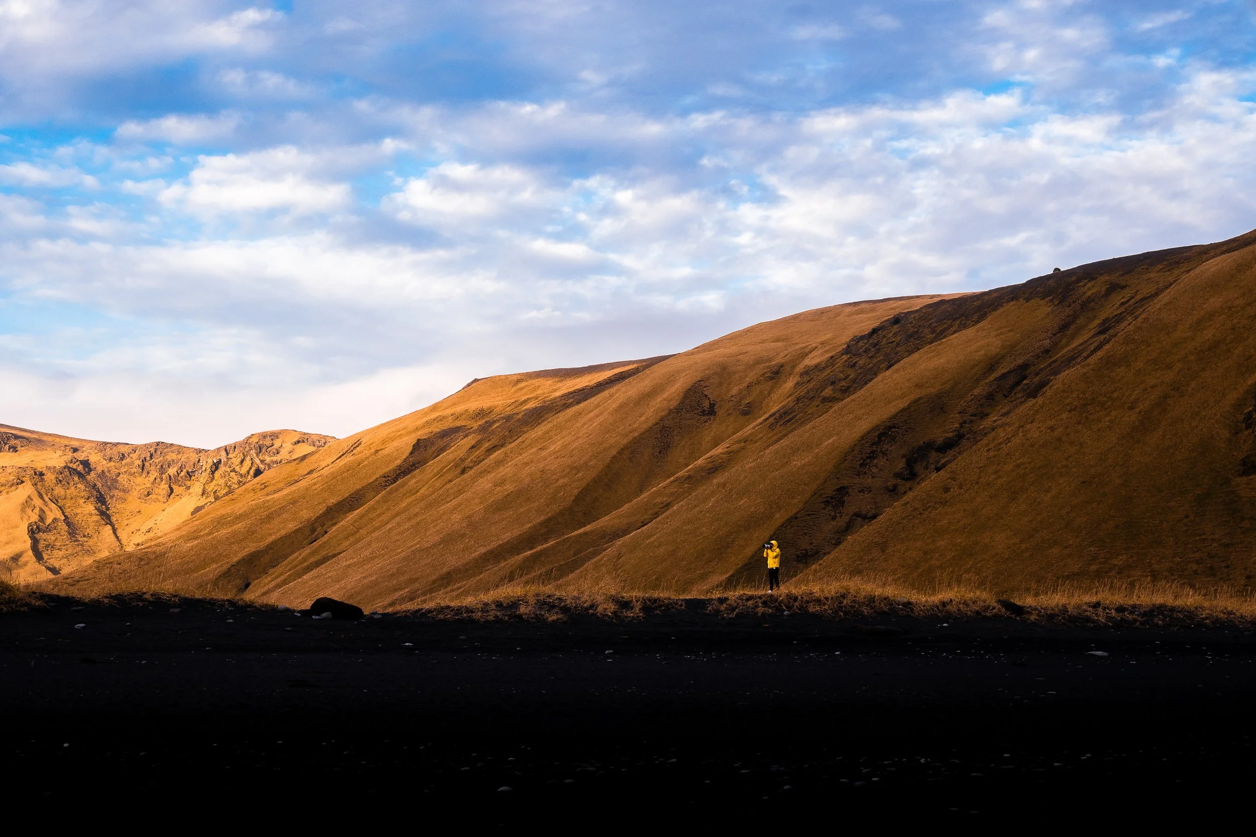 Hiker in a yellow jacket standing before sunlit brown mountain slopes beneath a cloudy blue sky