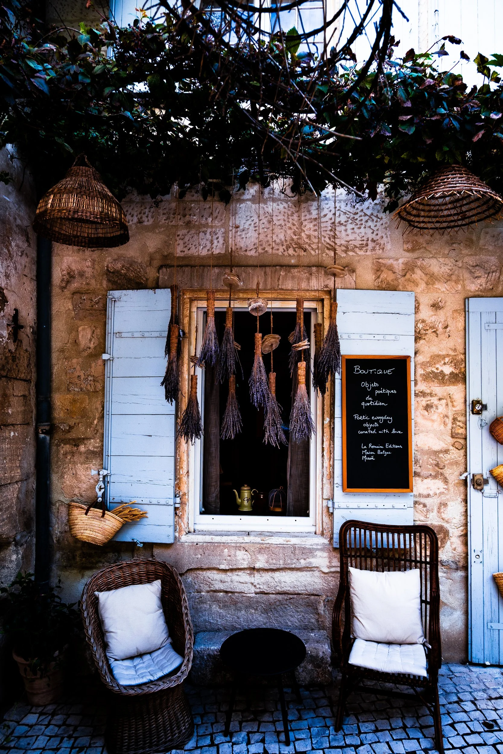 Rustic stone boutique storefront with blue shutters, hanging woven baskets, lavender bundles, and wicker chairs on a cobblestone street
