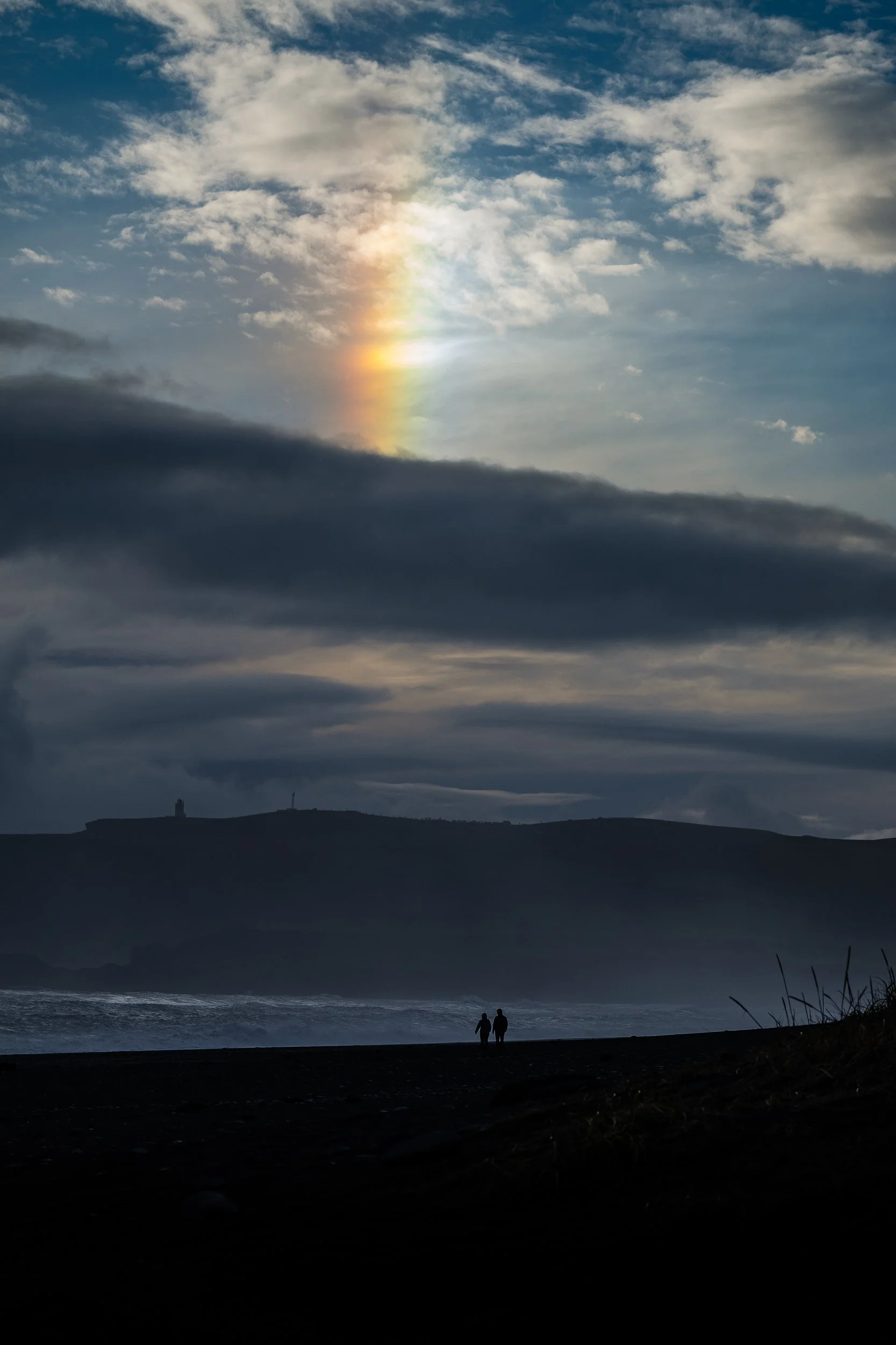 Silhouetted couple standing on a dark beach beneath storm clouds, with sunlight creating a bright rainbow-like halo over the ocean.