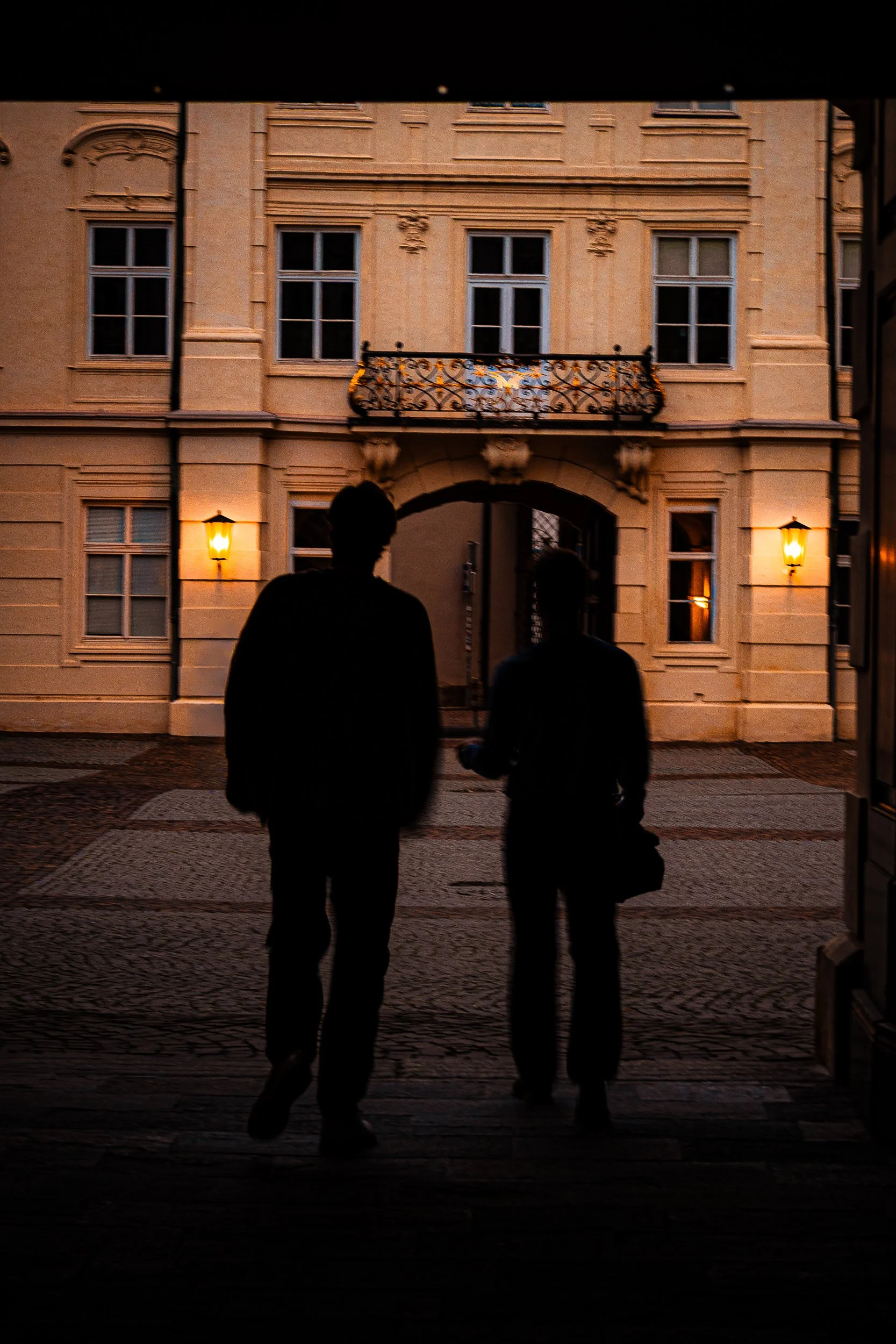 Two silhouetted people walking across a cobblestone courtyard at dusk in front of an ornate historic building with a wrought-iron balcony and glowing wall lanterns.
