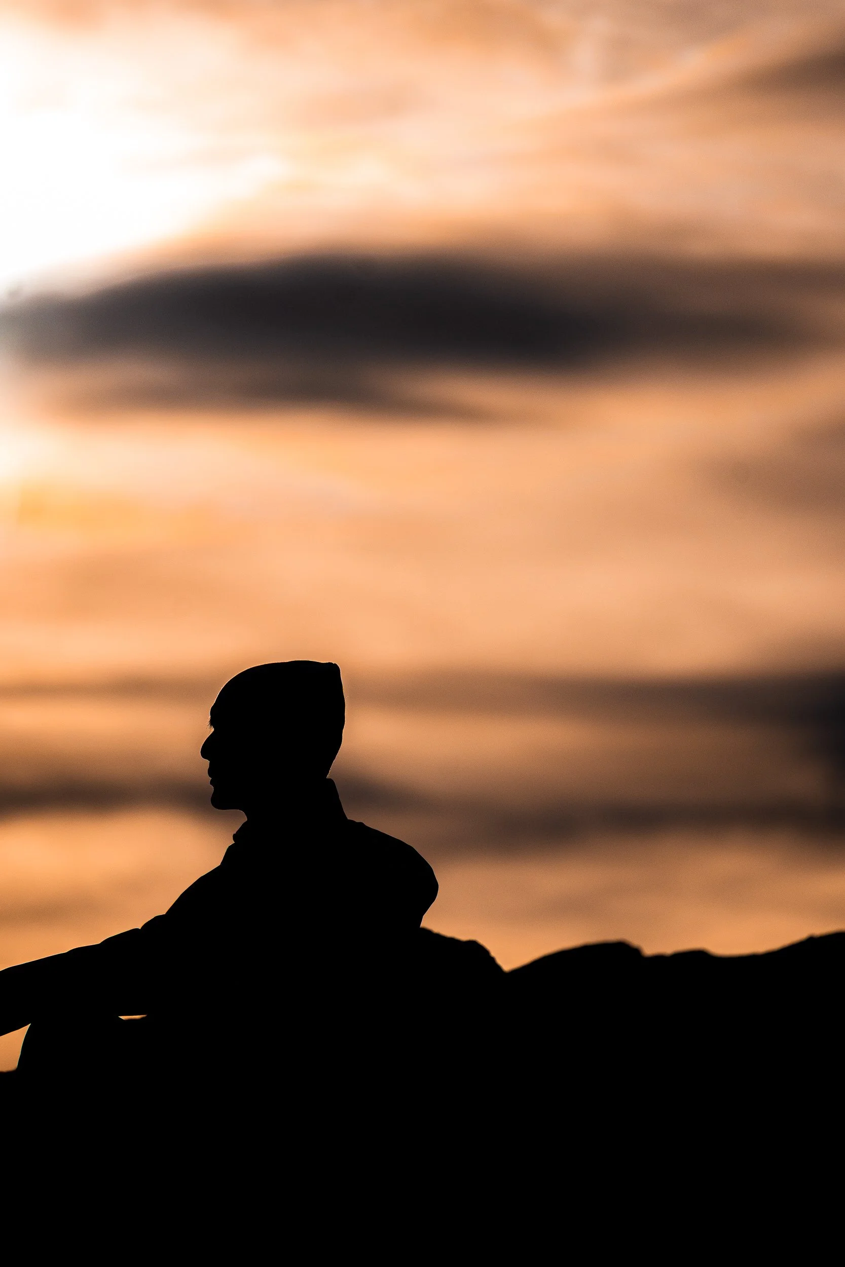 Silhouette of a seated person against a dramatic orange sunset sky and dark mountain landscape
