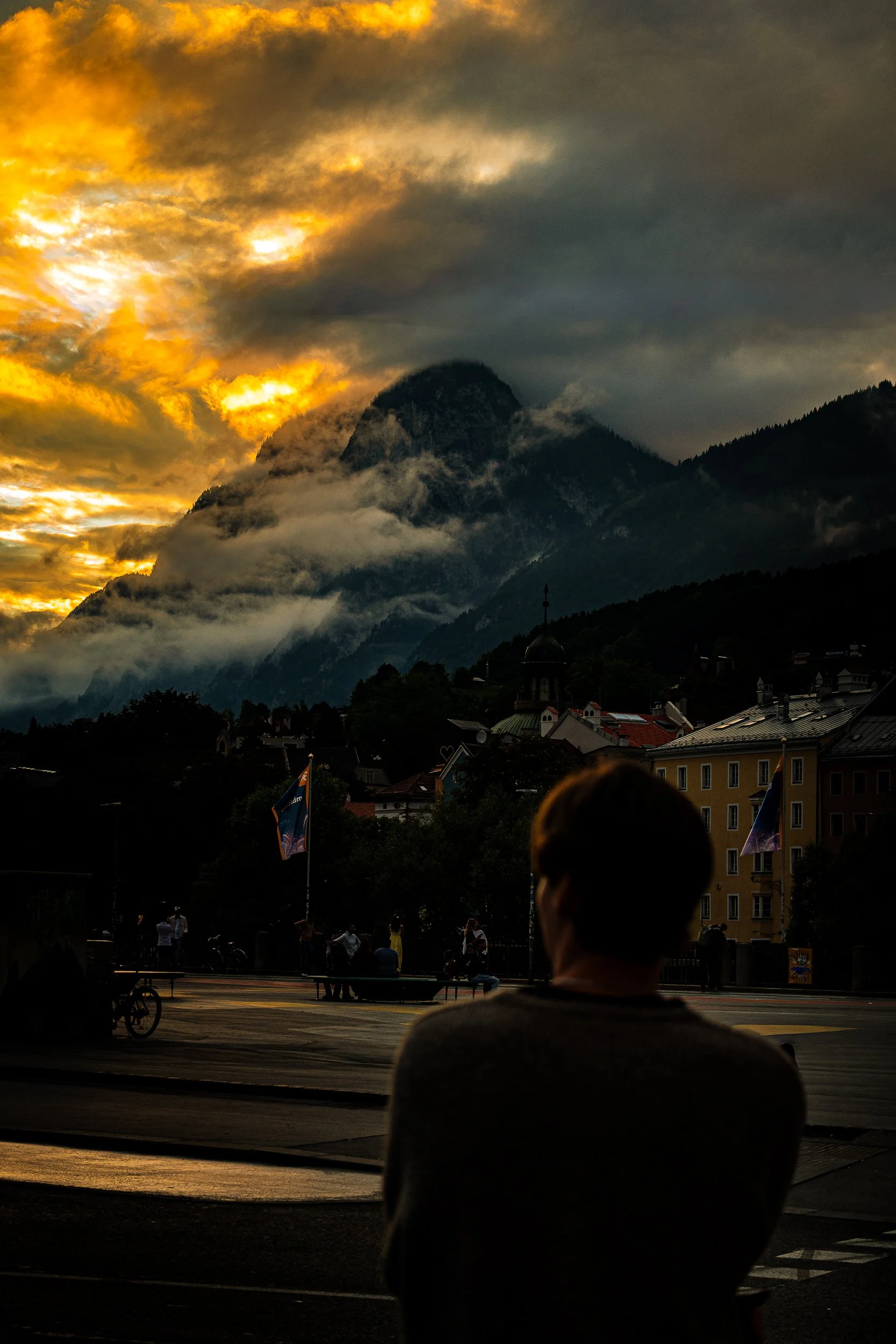 Dramatic golden sunset over misty Alpine mountains and a European town, with a silhouetted person in the foreground.