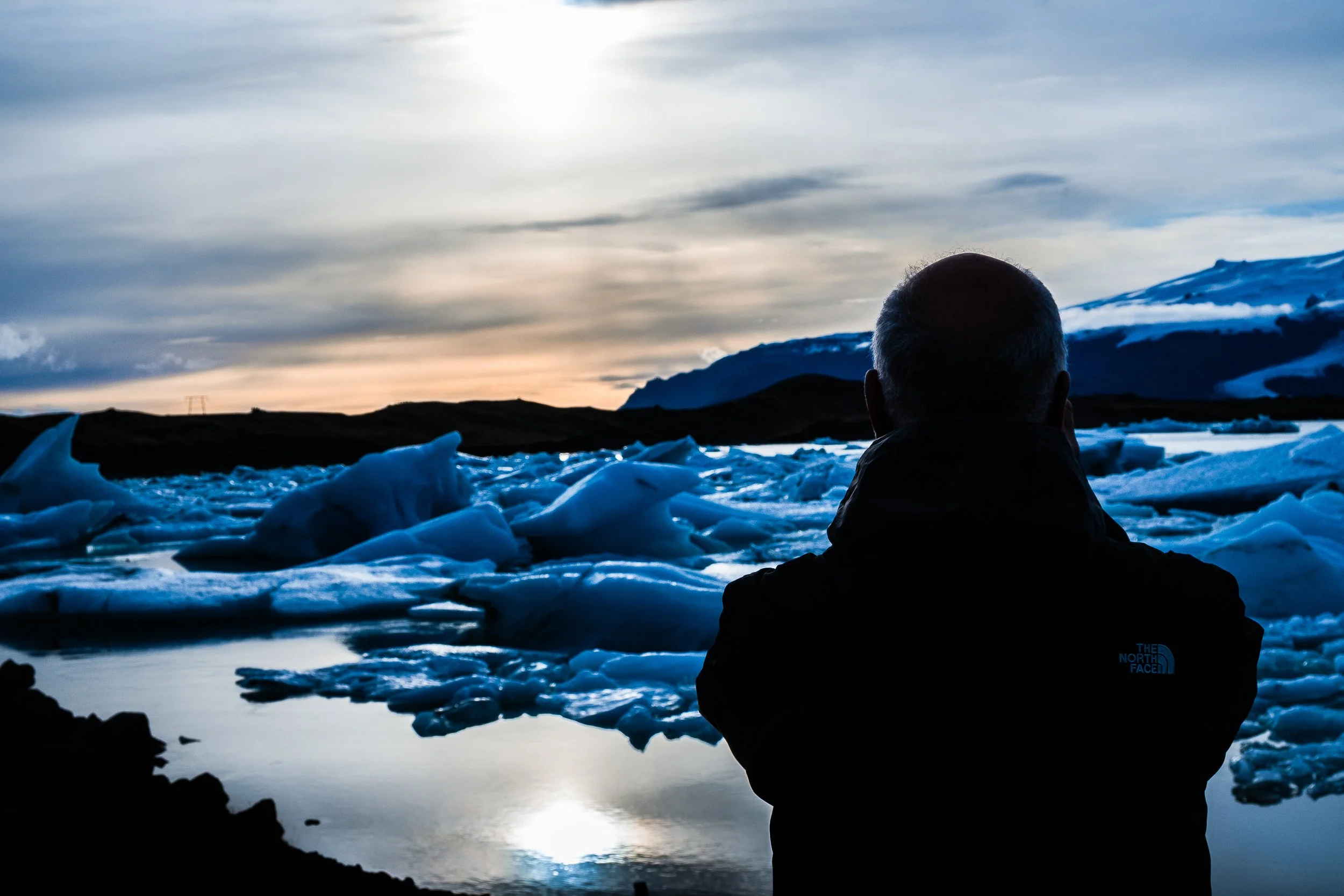 Person in a black jacket overlooking an iceberg-filled lagoon beneath snowy mountains at sunset