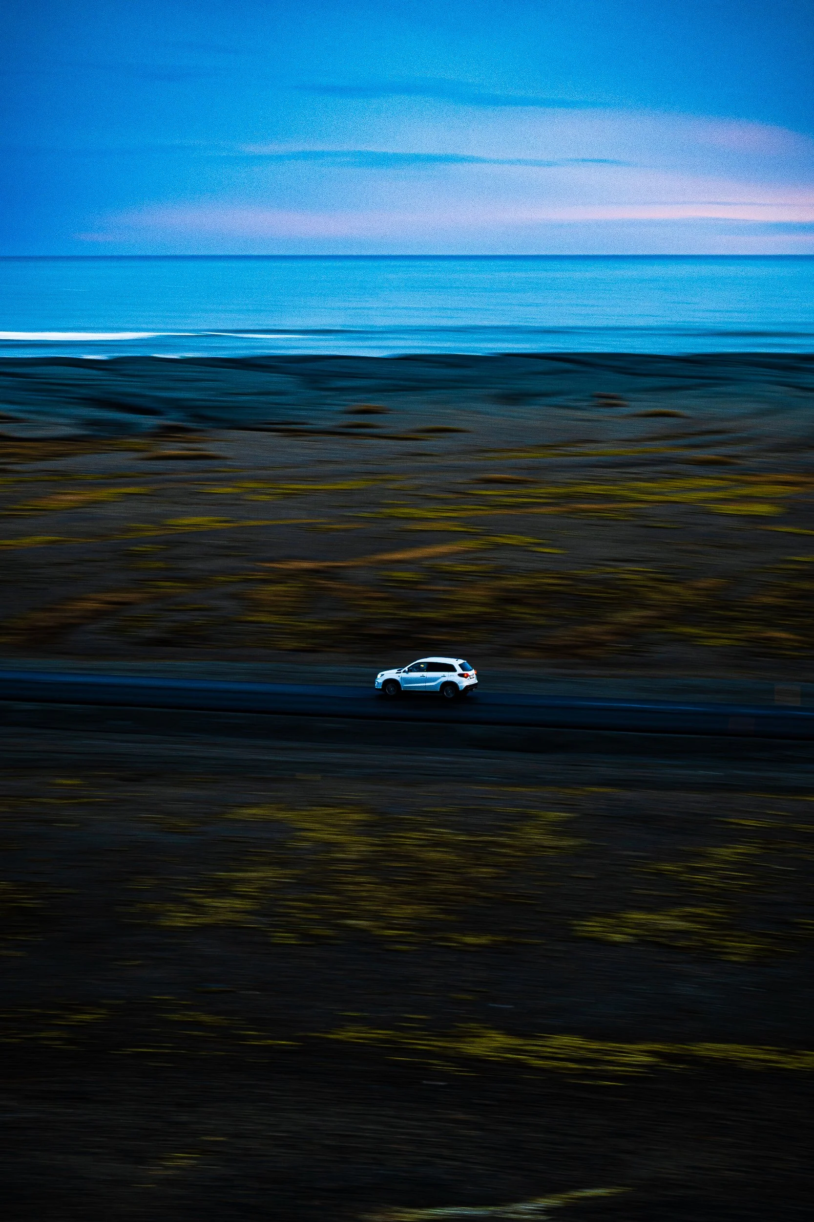 White SUV driving along a coastal road beside the ocean at dusk