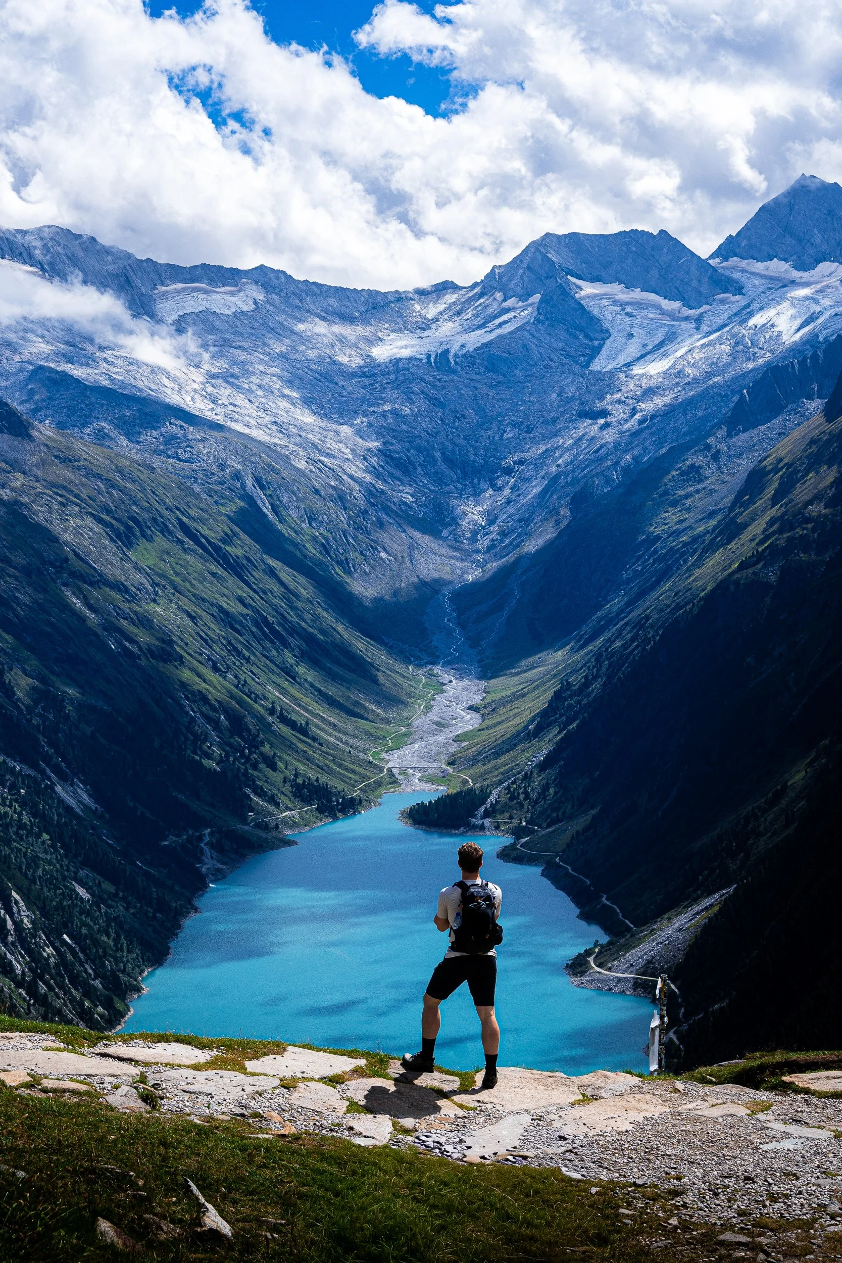 Hiker overlooking a turquoise alpine lake, glacier-carved valley, and snow-capped mountains beneath a cloudy blue sky