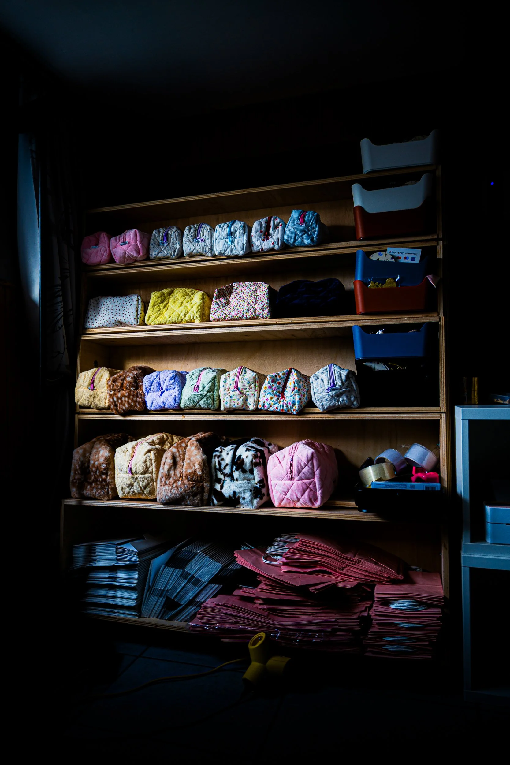 Wooden shelving unit stocked with colorful quilted fabric pouches, storage bins, and stacks of paper bags