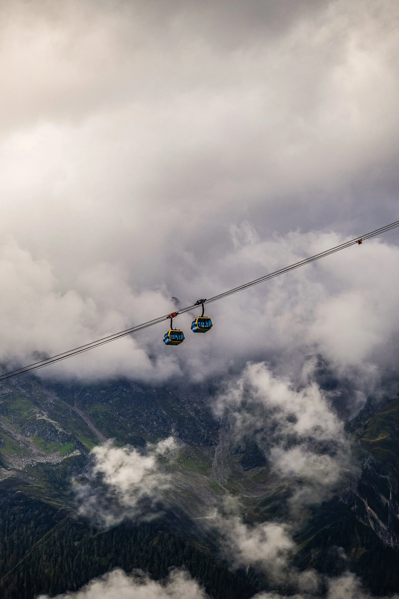Two blue-and-yellow cable cars suspended over a rugged, cloud-covered mountain valley