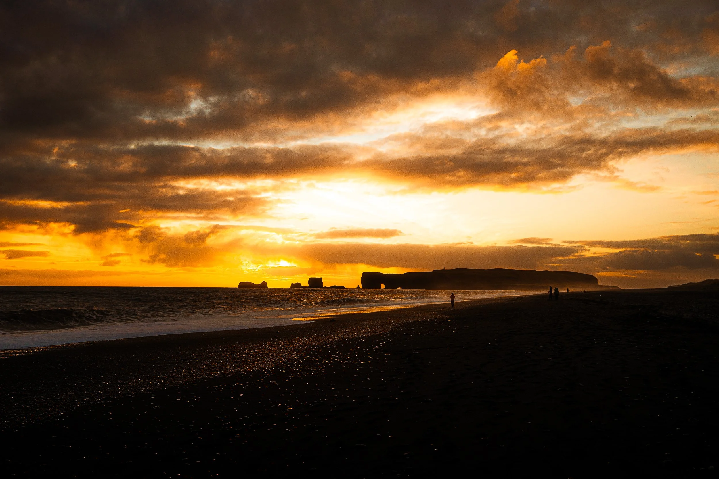 Sunset over a black-sand beach with ocean waves, dramatic clouds, and offshore rock formations