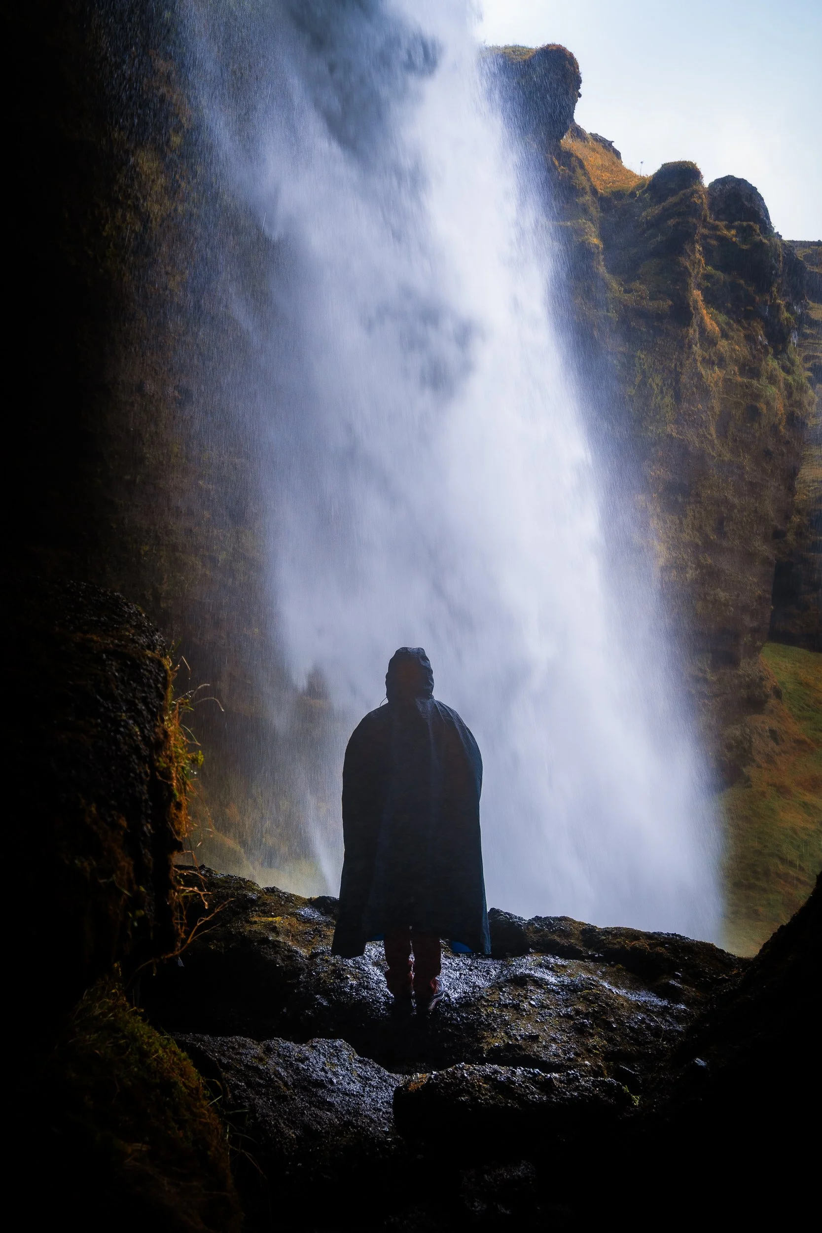 Hiker in a dark rain poncho standing beneath a powerful waterfall surrounded by mossy cliffs