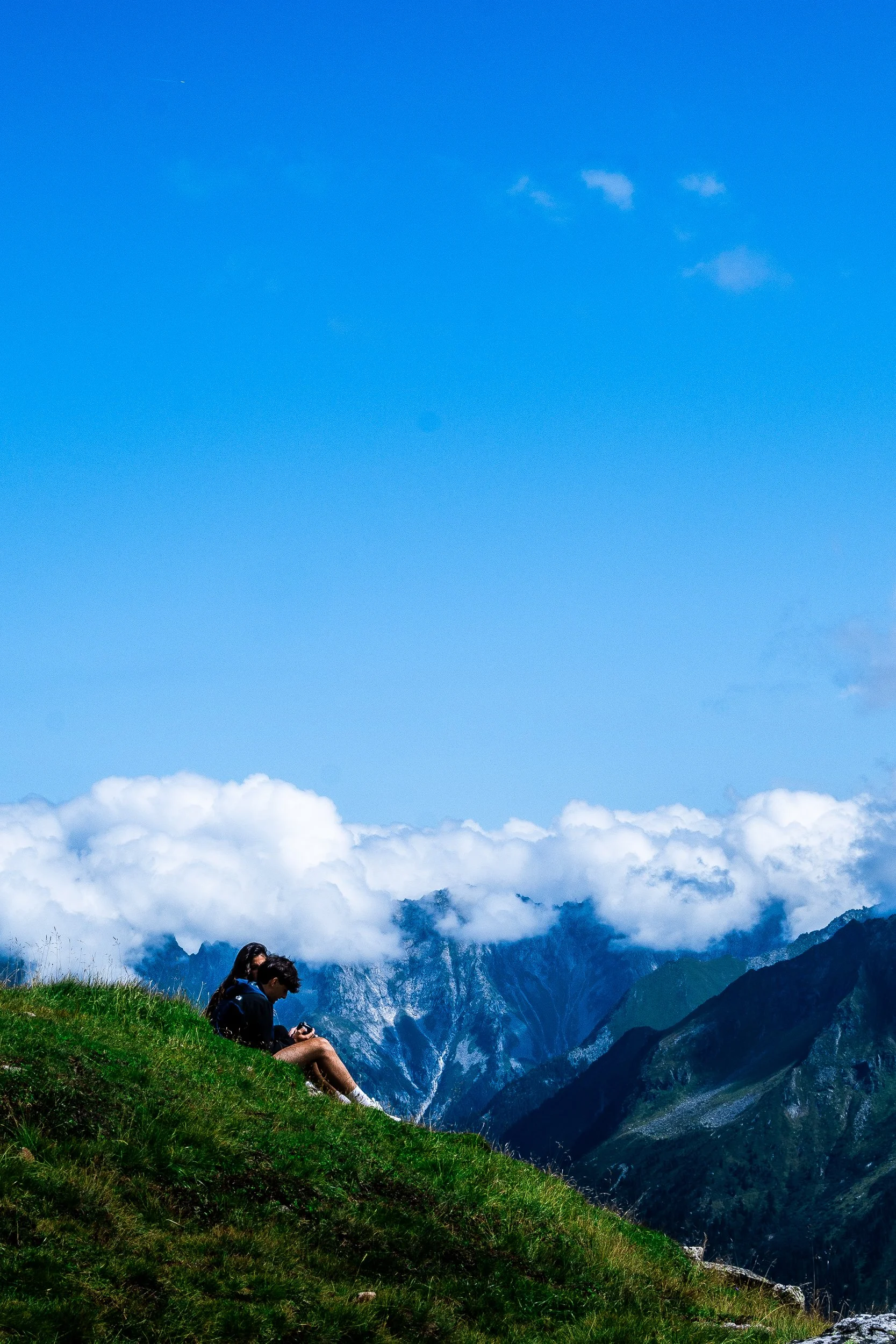 Two hikers sitting on a grassy mountain slope overlooking cloud-covered alpine mountains beneath a bright blue sky