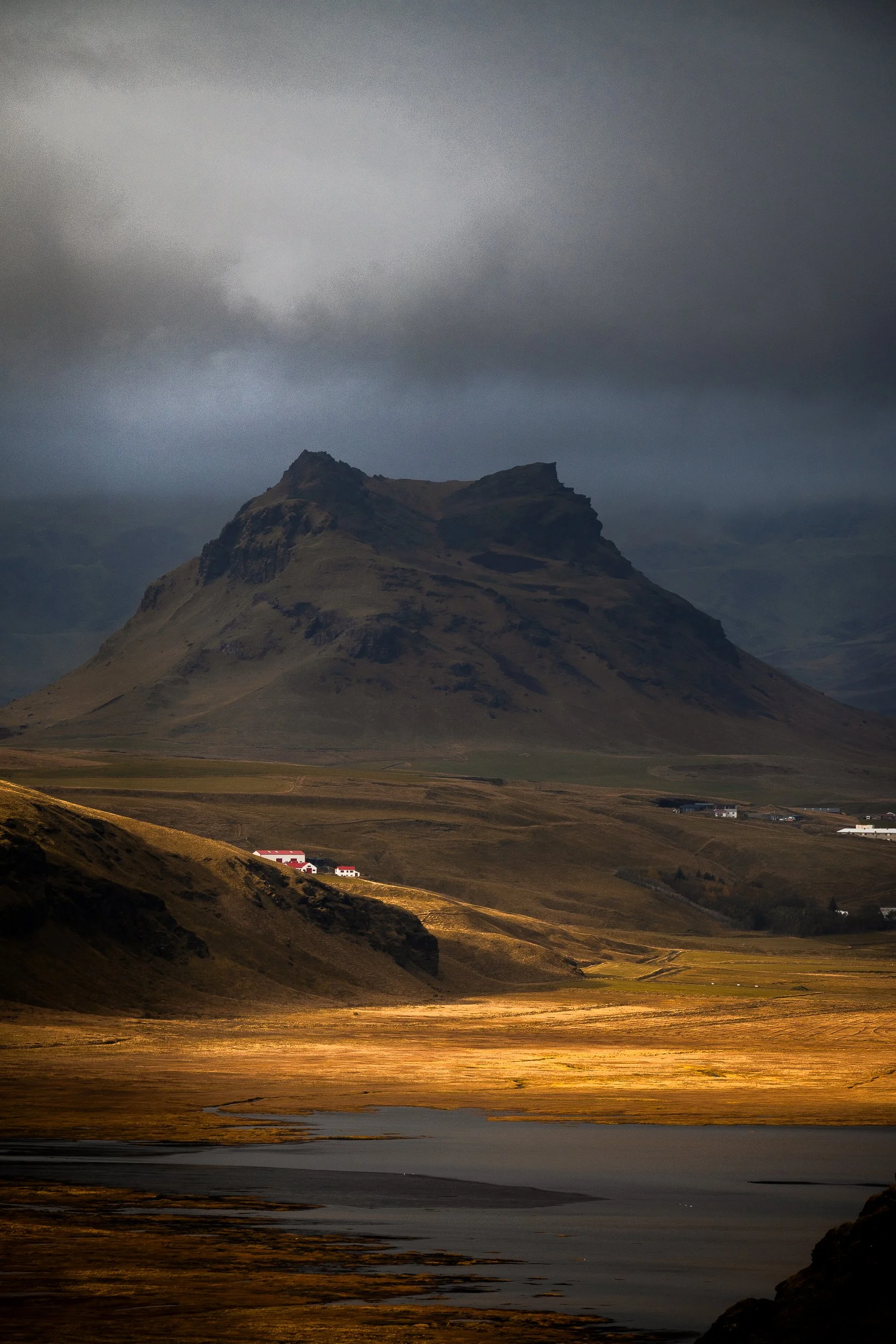 Dramatic Icelandic mountain landscape with a dark volcanic peak, golden fields, scattered white houses, and a calm lake under storm clouds