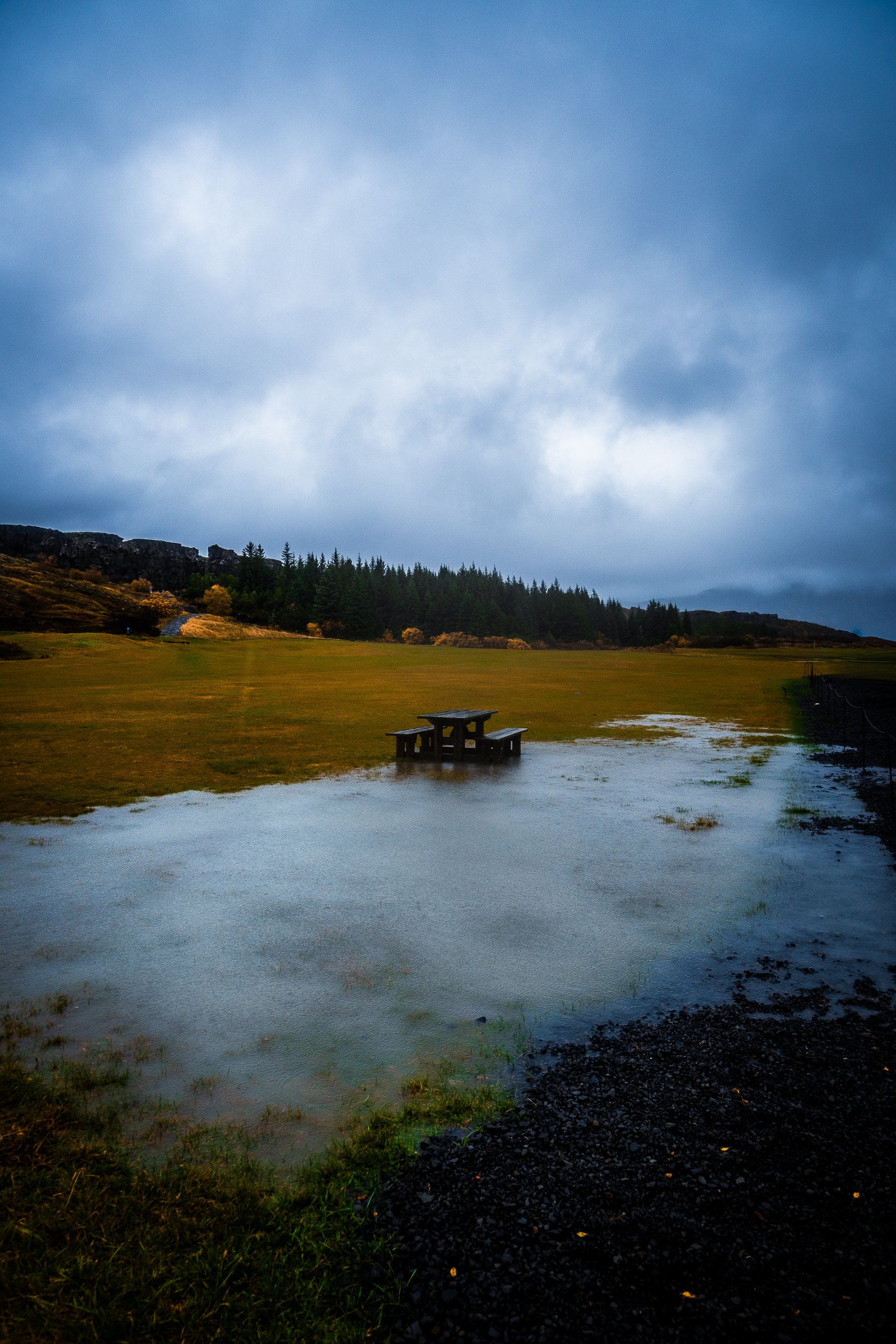 Rain-soaked picnic table beside a flooded grassy field, evergreen forest, and rocky cliffs under a stormy sky