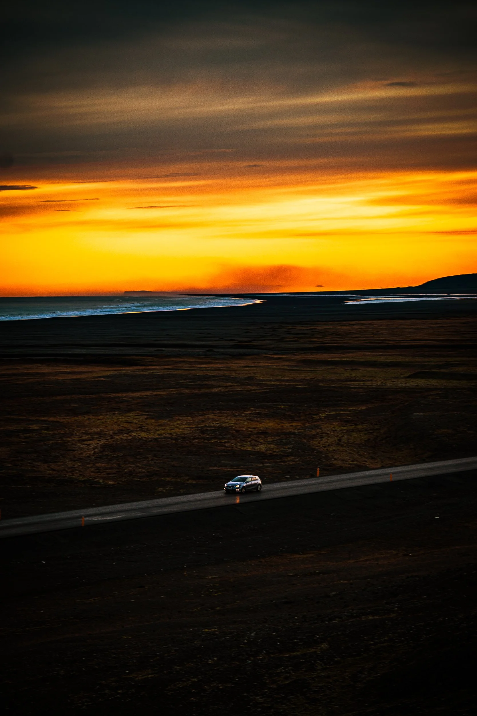 A car drives along a coastal road beside dark sand beaches and ocean waves at sunset beneath glowing orange clouds.