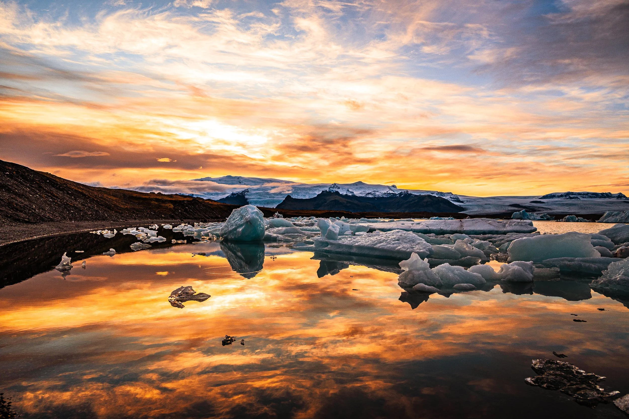 Sunset over an Icelandic glacier lagoon with floating icebergs, snow-covered mountains, and reflected orange clouds