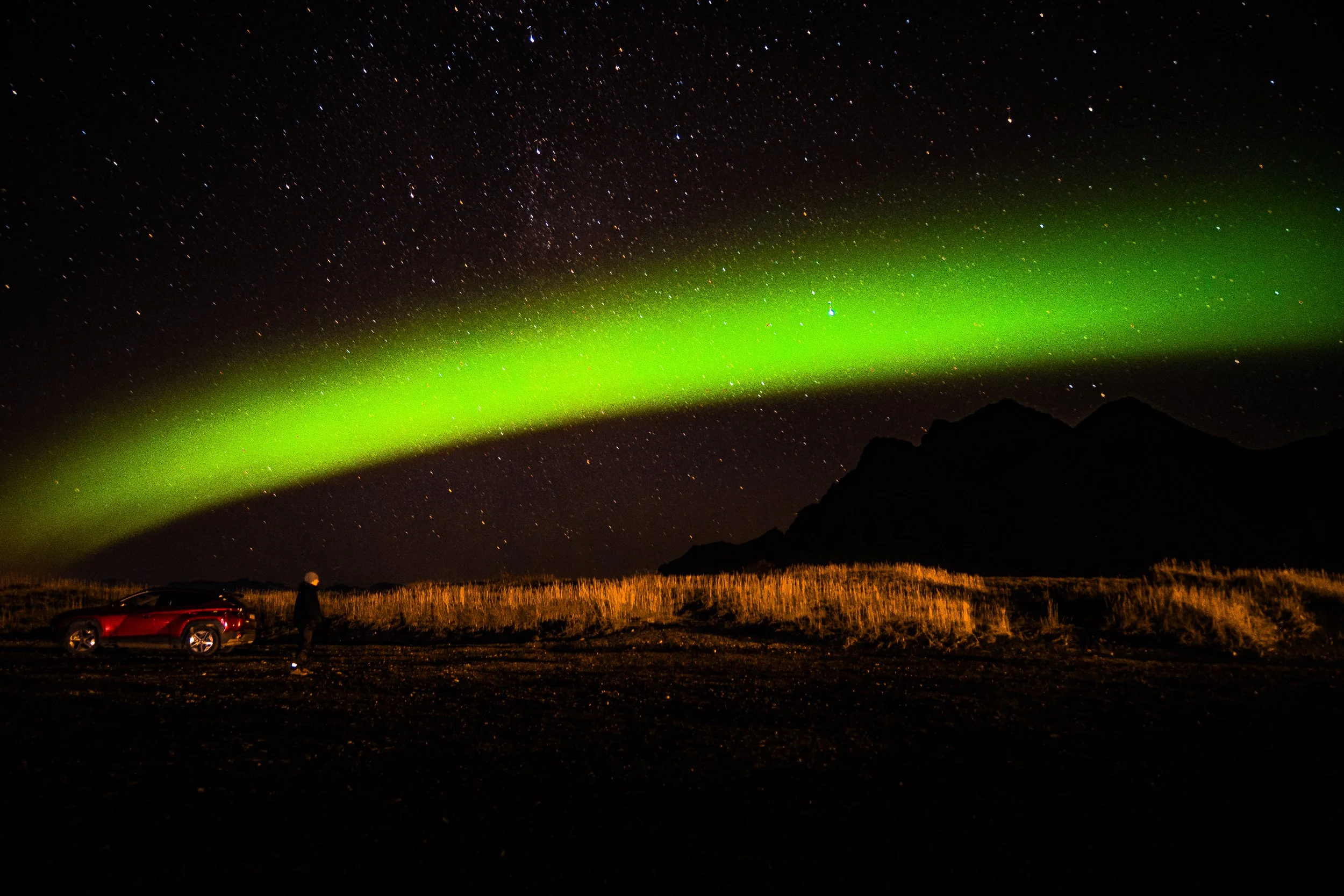 Green aurora borealis glowing above a starry night sky, silhouetted mountains, golden grass, and a red car with a person.