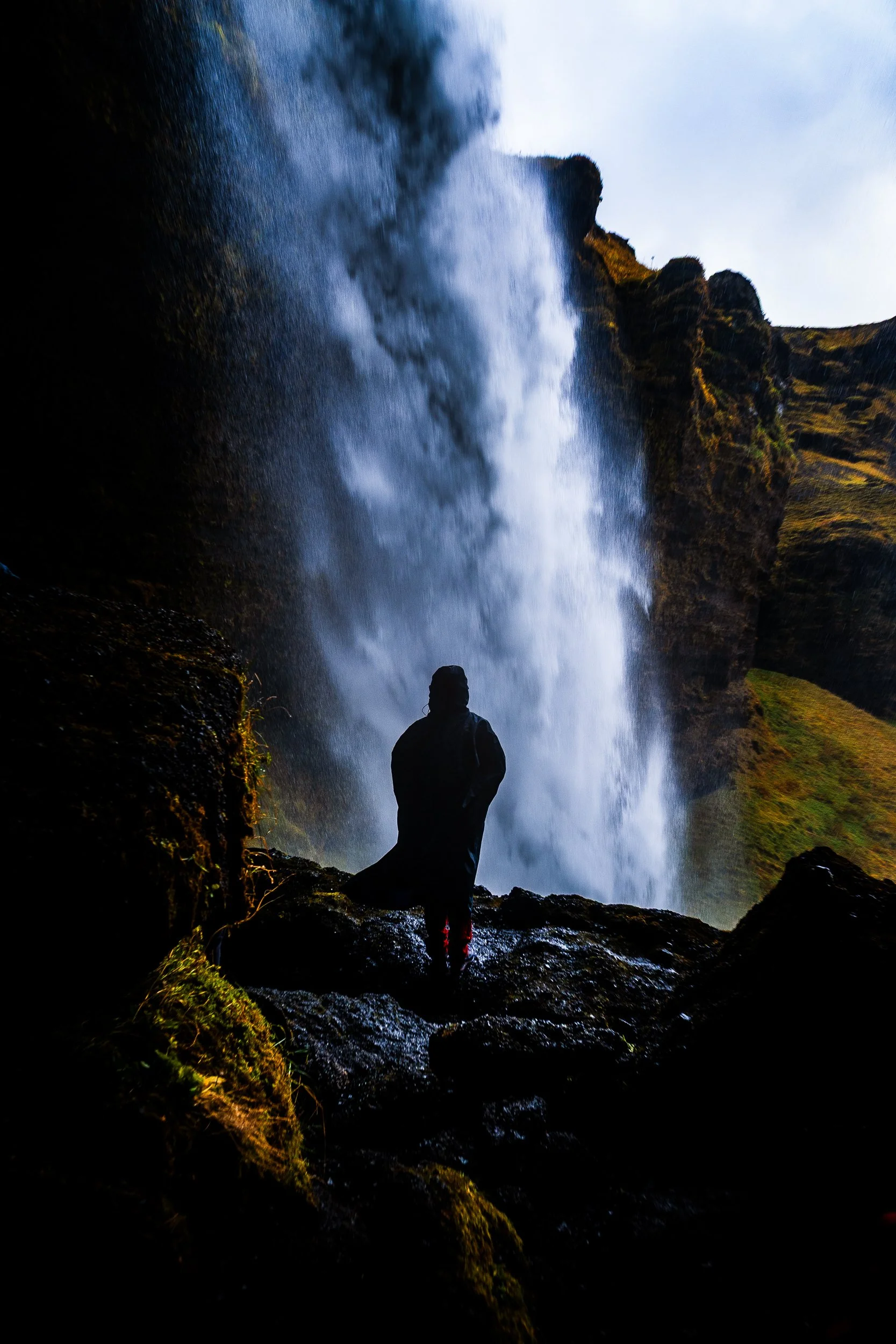 Silhouetted person standing beneath a powerful waterfall surrounded by mossy cliffs