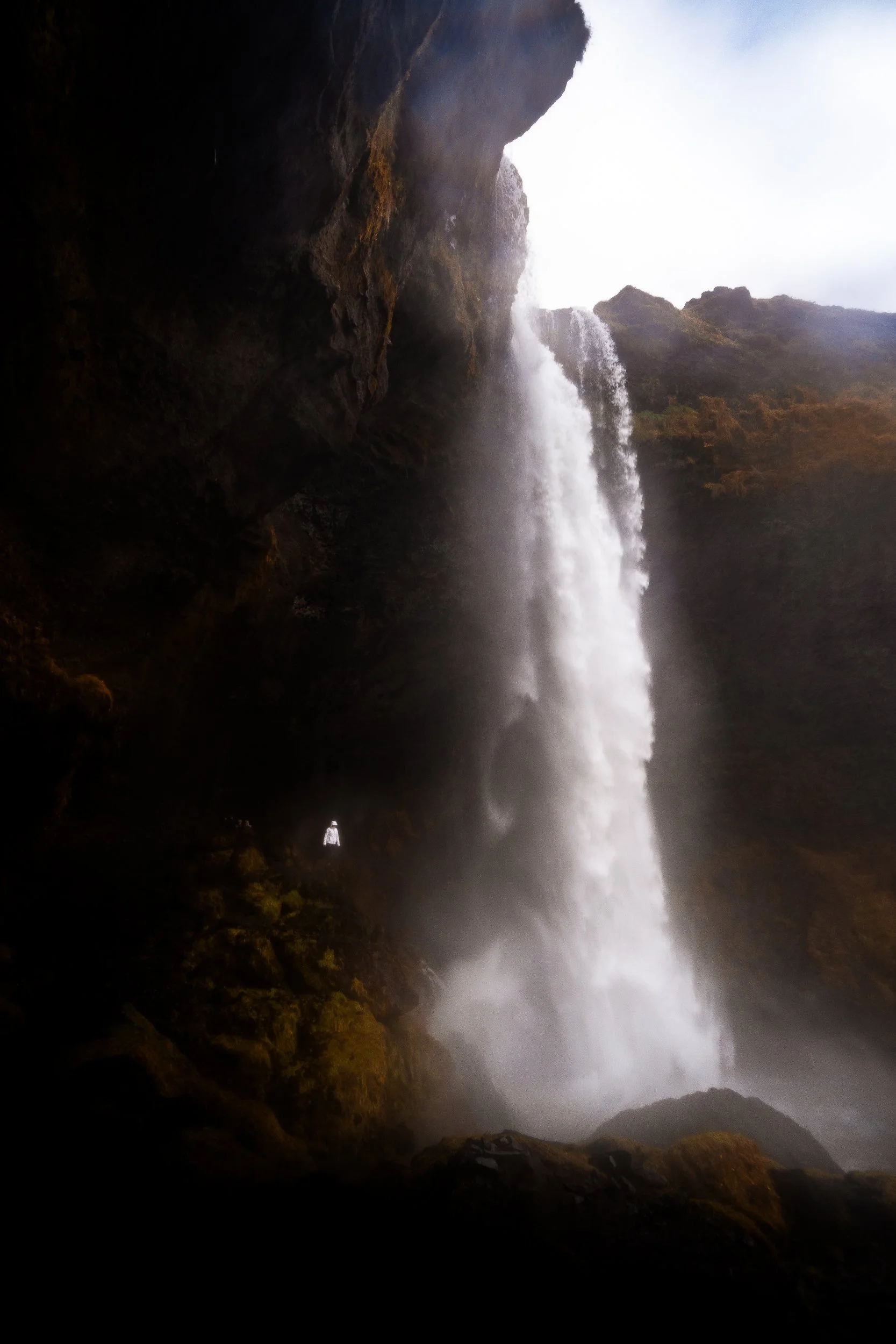 Tall waterfall cascading beside a dark moss-covered cliff, with a tiny person standing near its base.