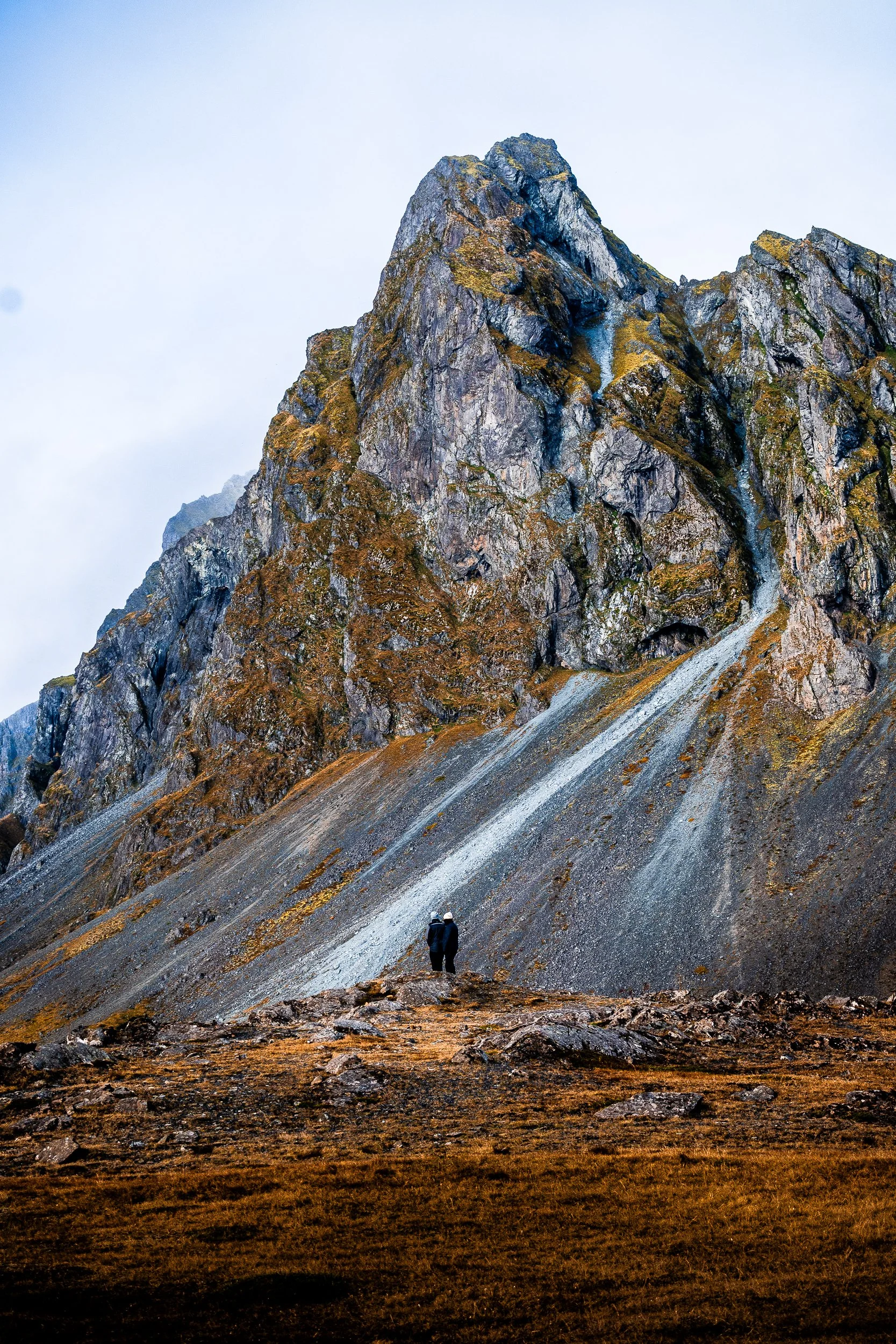 Hiker standing below a rugged, lichen-covered mountain peak and steep rocky slope