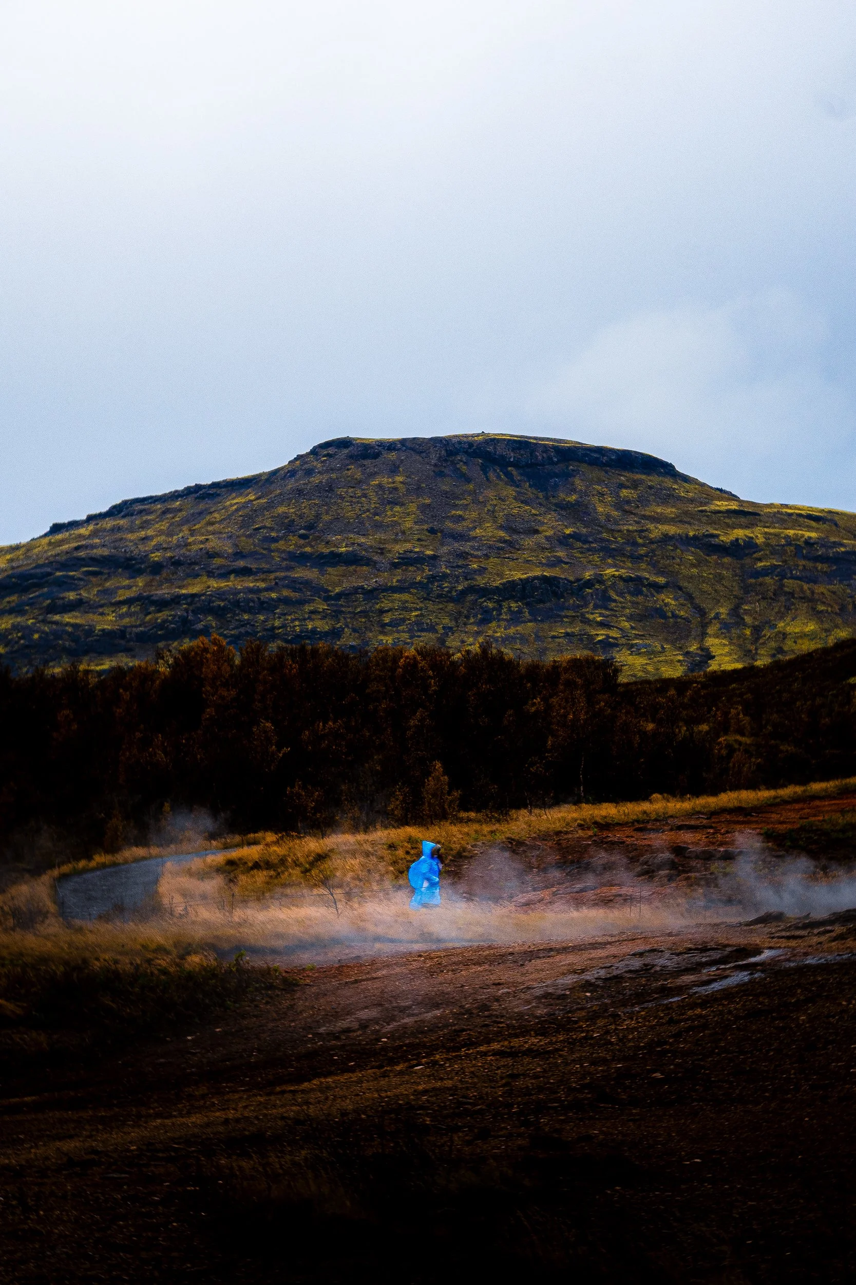 Person in a blue jacket walking through a steaming geothermal landscape beneath a moss-covered volcanic mountain