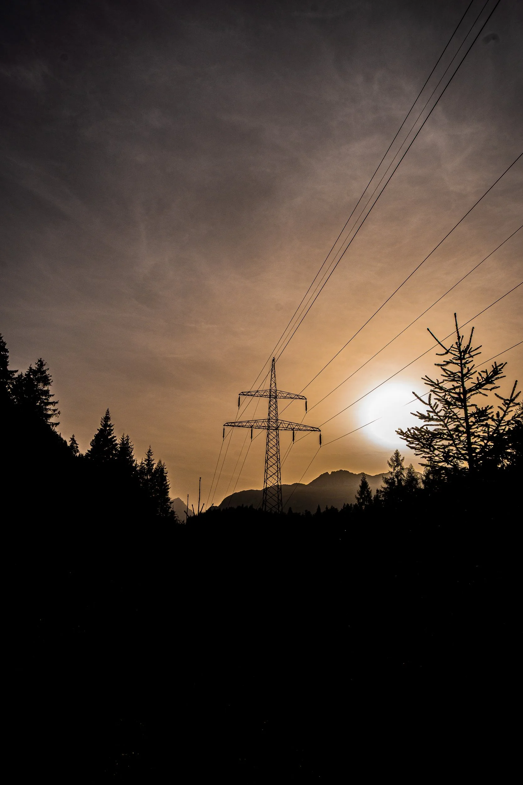 Silhouetted power transmission tower and lines against a sunset over evergreen trees and mountain peaks