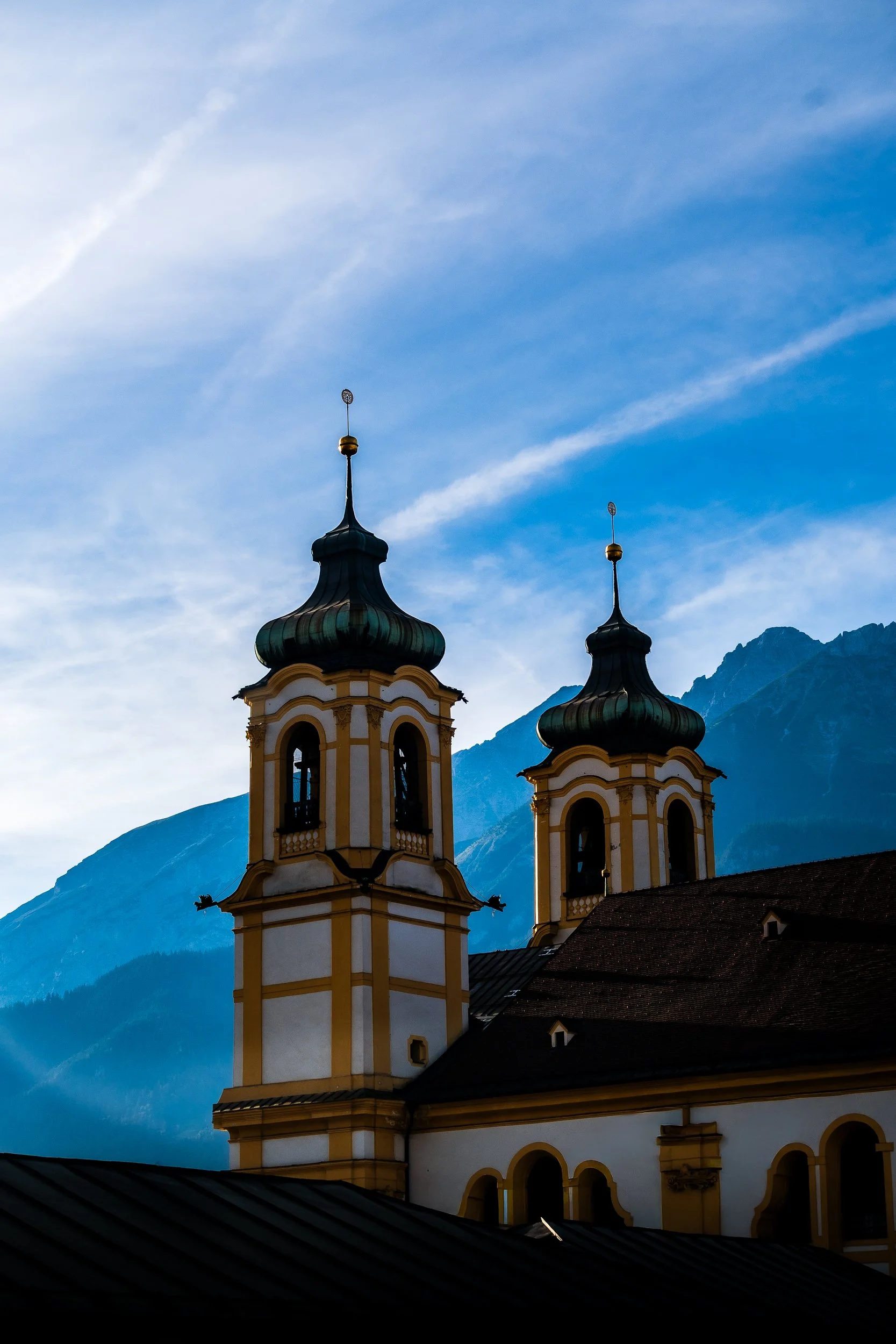 Baroque church towers with dark onion domes against a blue sky and rugged Alpine mountains