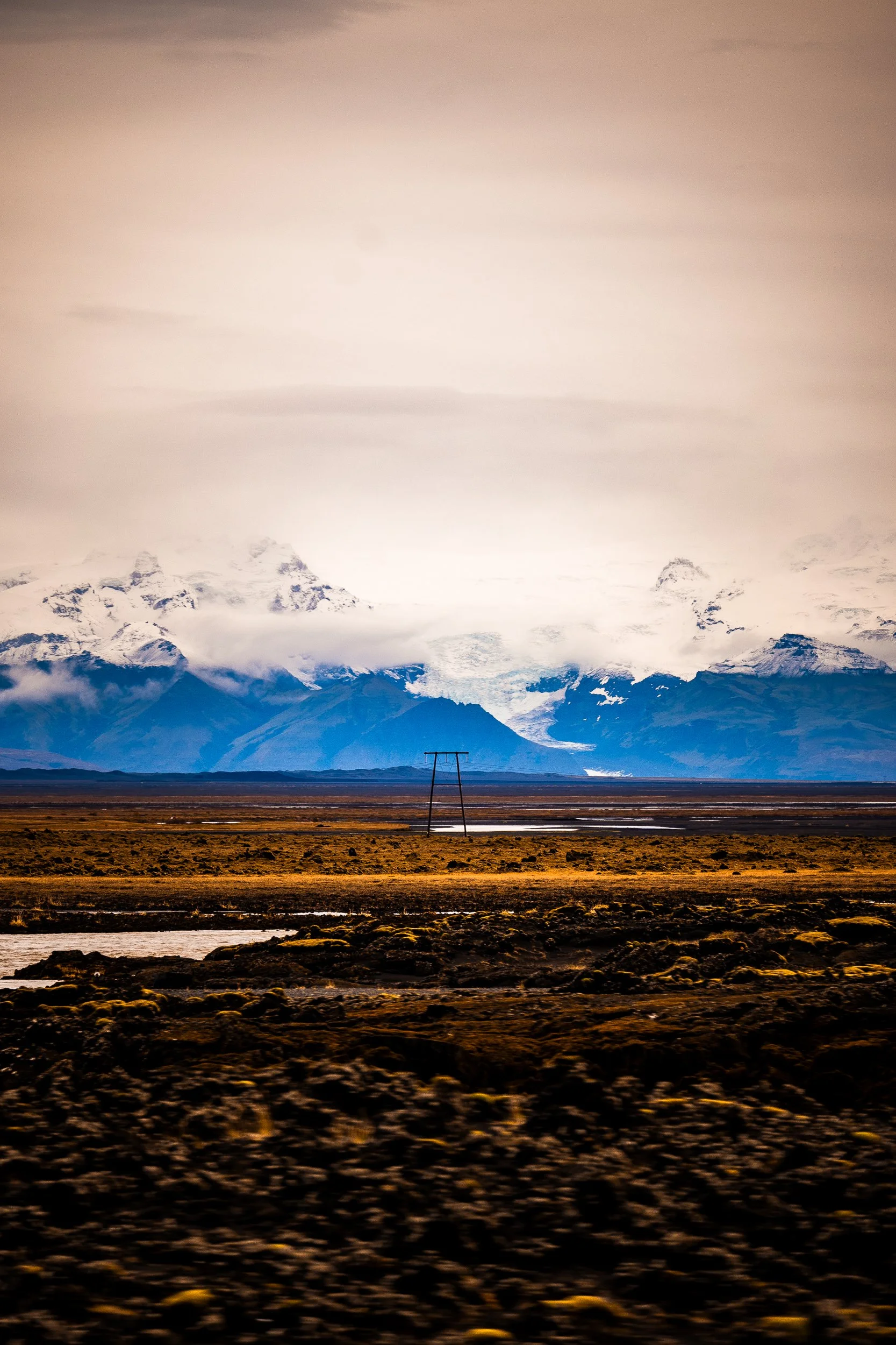 Snow-covered mountain range beneath clouds, viewed across an Icelandic mossy plain with shallow water and utility poles.