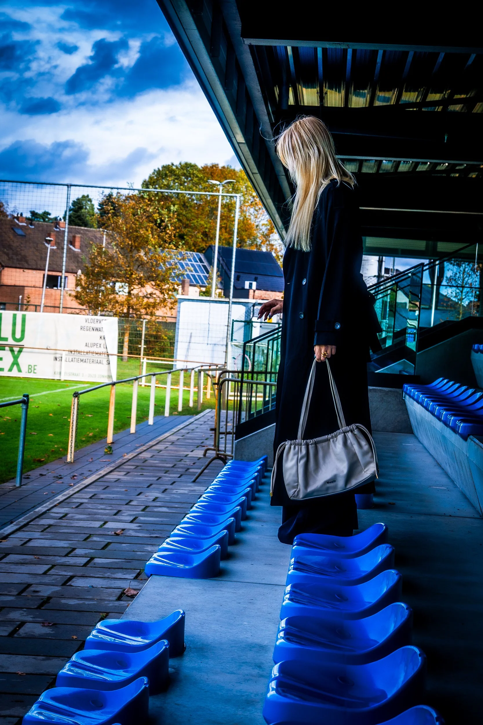 Blonde woman carrying a handbag through blue stadium seats beside an outdoor sports field.