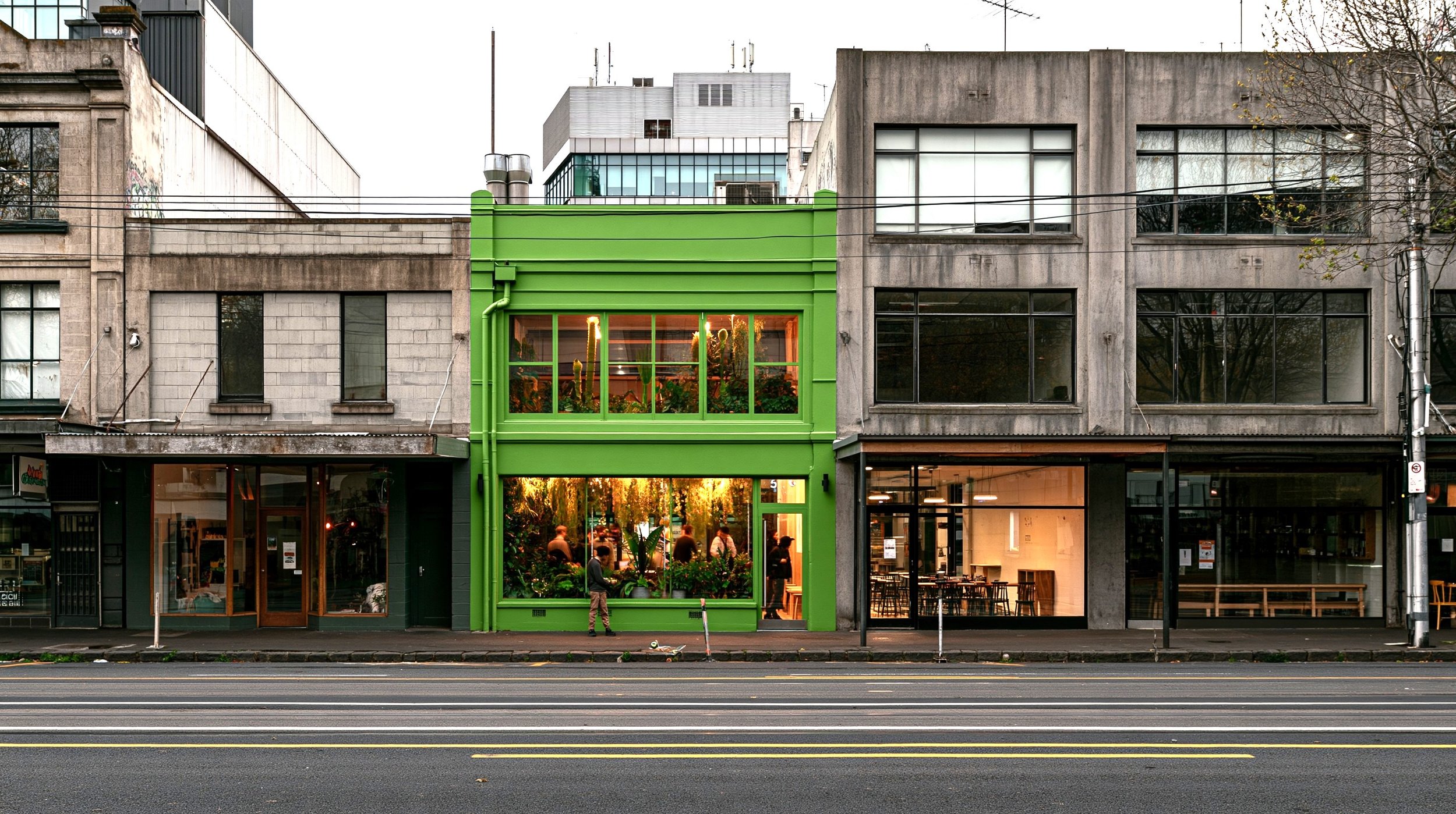 A city street with a vibrant green storefront, large windows revealing a lush indoor plant-filled space, and adjacent of older gray and beige buildings, with a few pedestrians and cars.