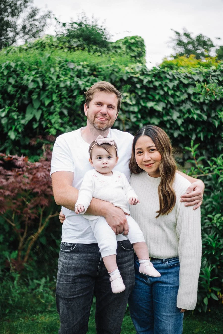 Husband and wife and their baby standing in a garden smiling.