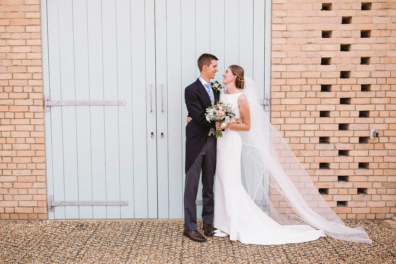 Newlywed bride and groom smiling together outside a brick building, with the bride holding a bouquet and wearing a long veil.