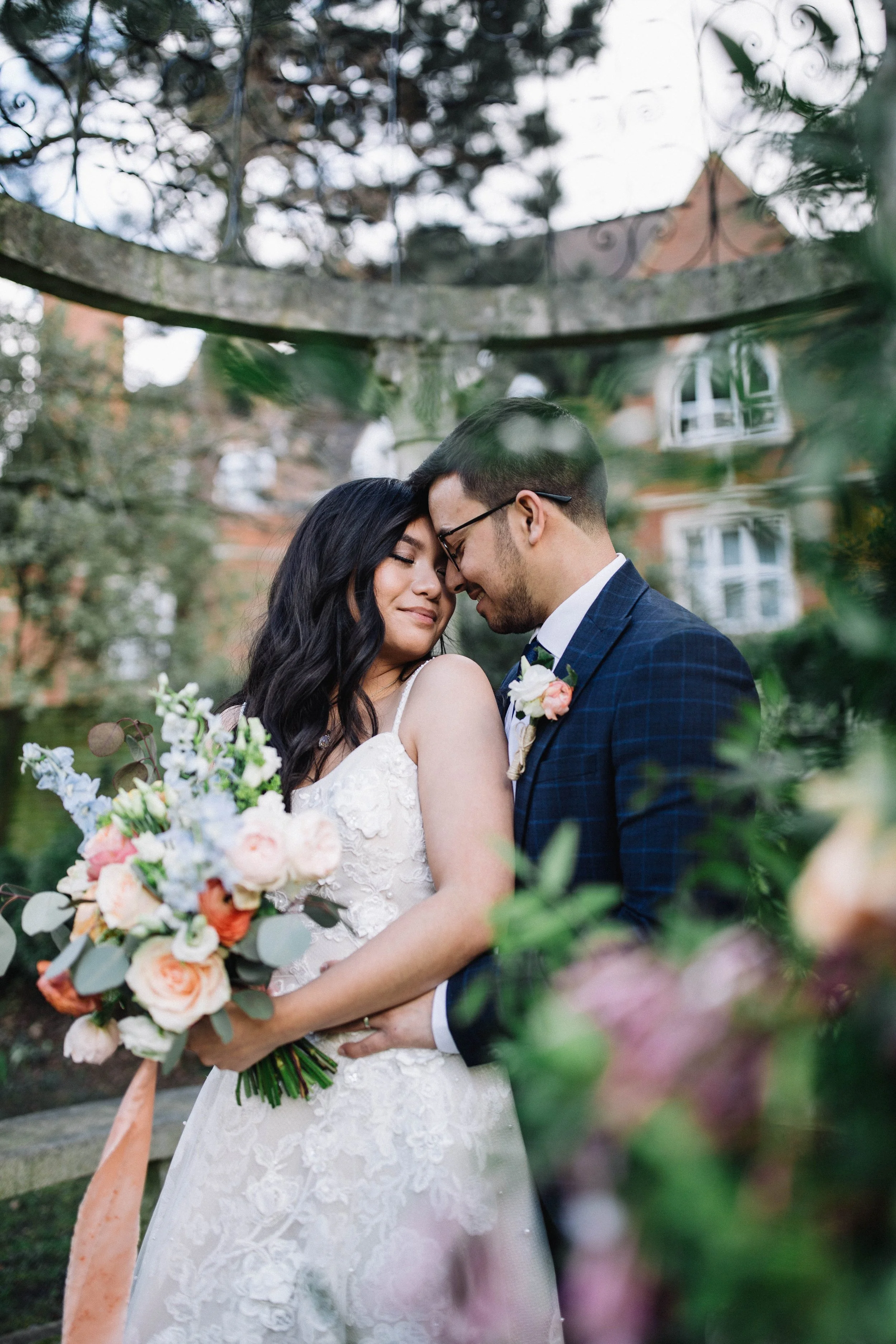 The bride holds a wedding bouquet and leans back towards the groom, their foreheads are touching and eyes are closed.