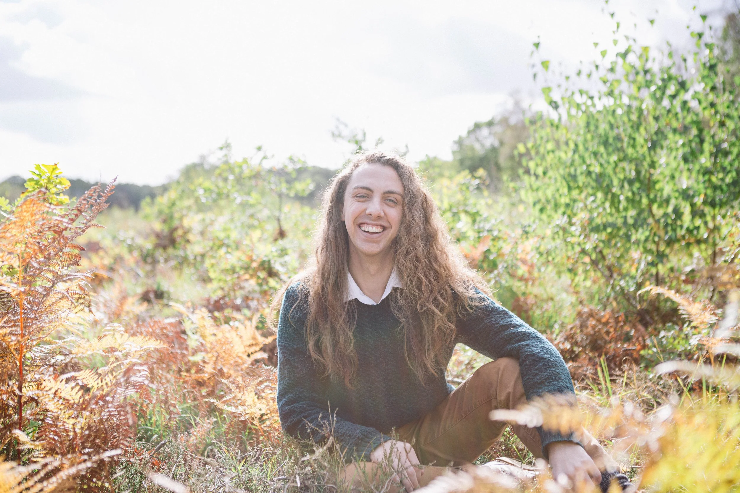 Smiling man with long hair sitting outdoors among lush greenery and autumn foliage