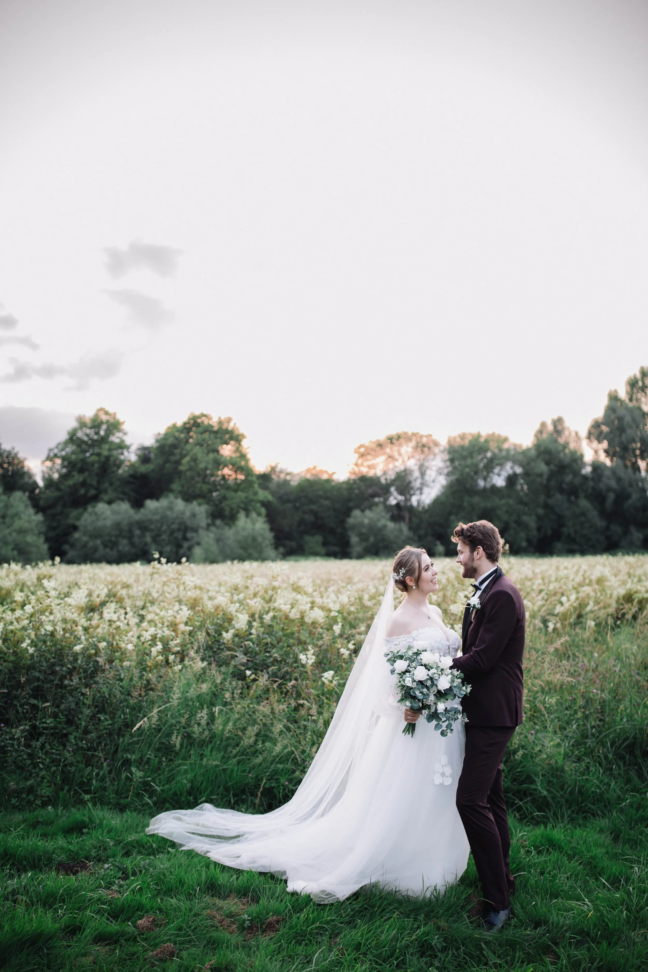 Bride and groom embrace, facing each other. The sunset just behind them over the flowers in the field.