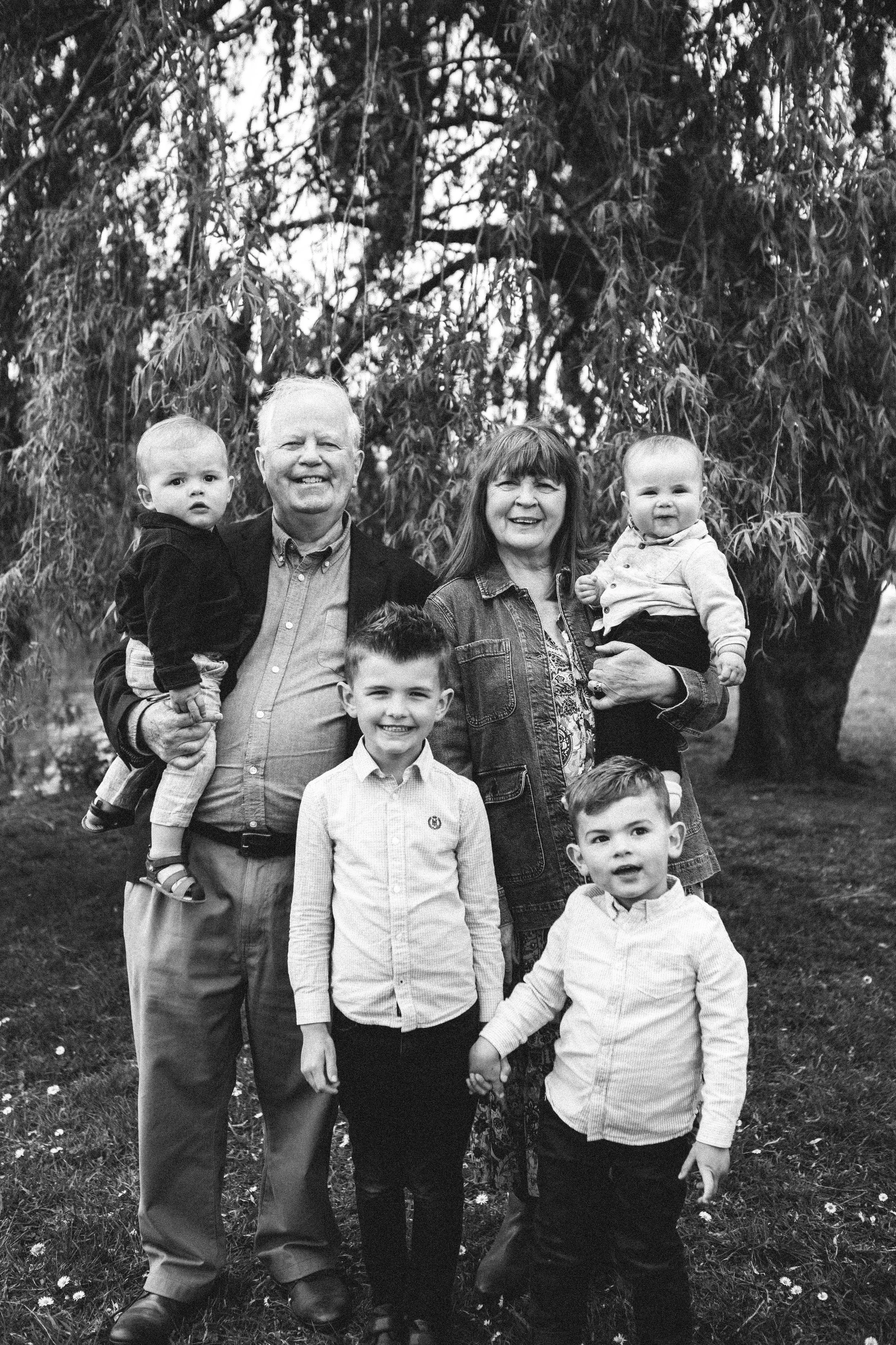 Black and white of grandparents holding there grandsons. With two more grandsons standing in front of them smiling under a willow tree. 