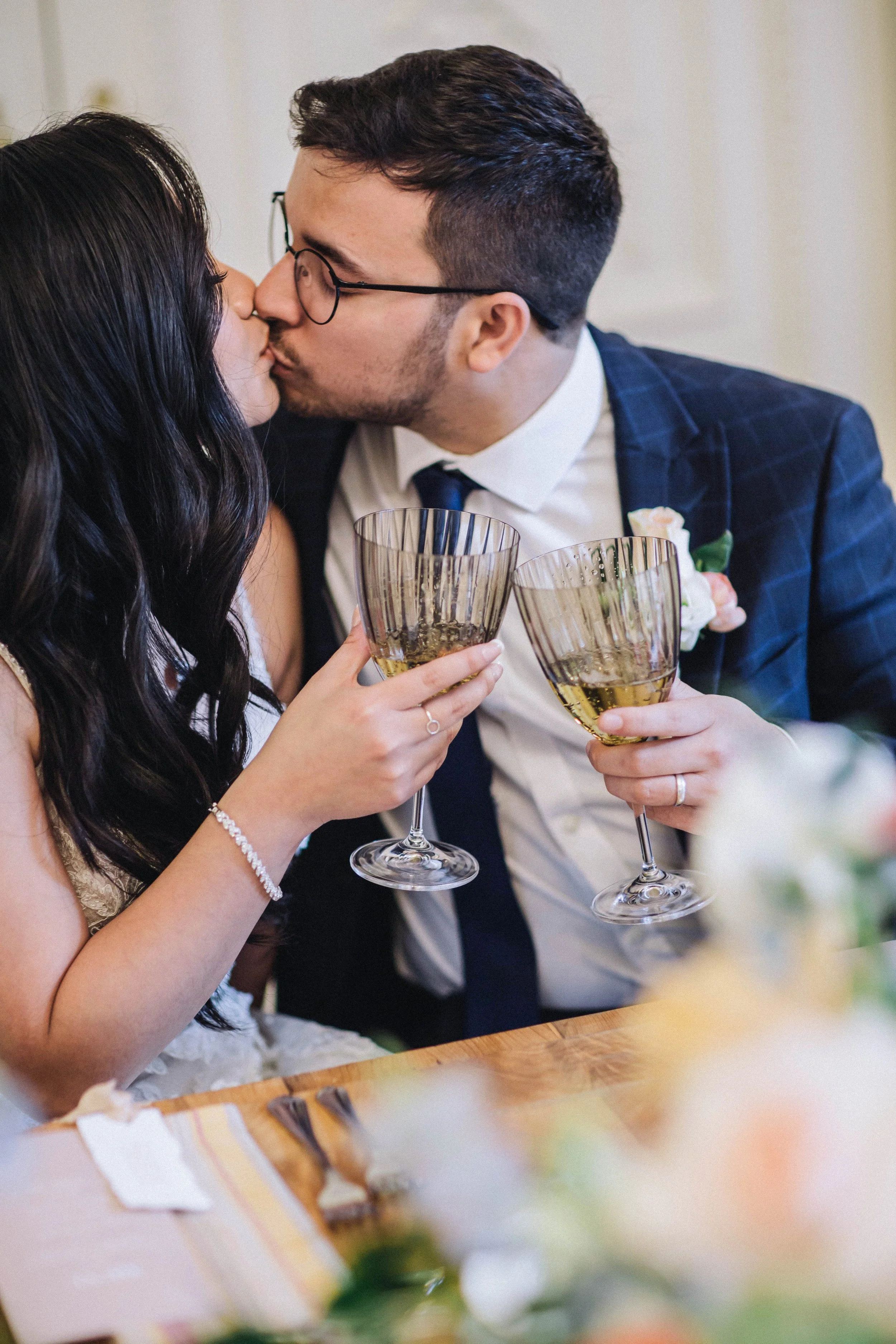 Bride and groom cheers as they kiss, sitting at their wedding table.