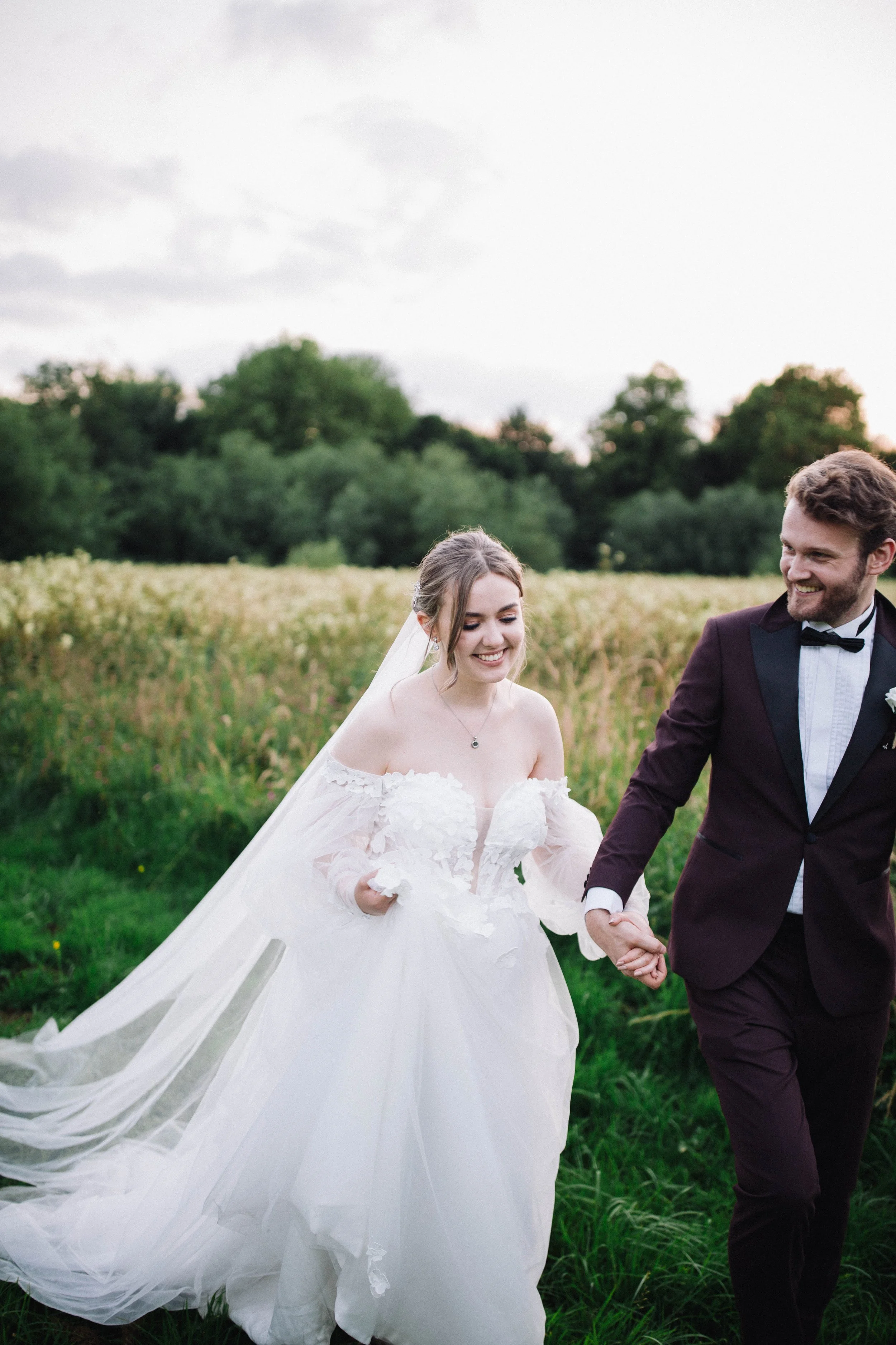 Groom leading bride through the grassy field while both are smiling.