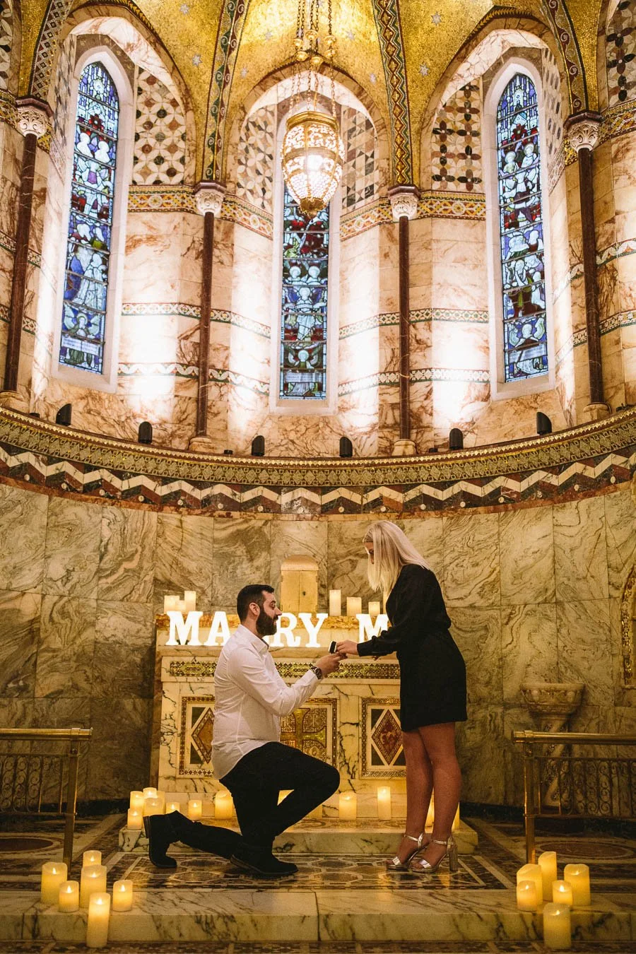 Man down on one knee to propose inside Fitzrovia Chapel. Woman looks down at him excitedly smiling.