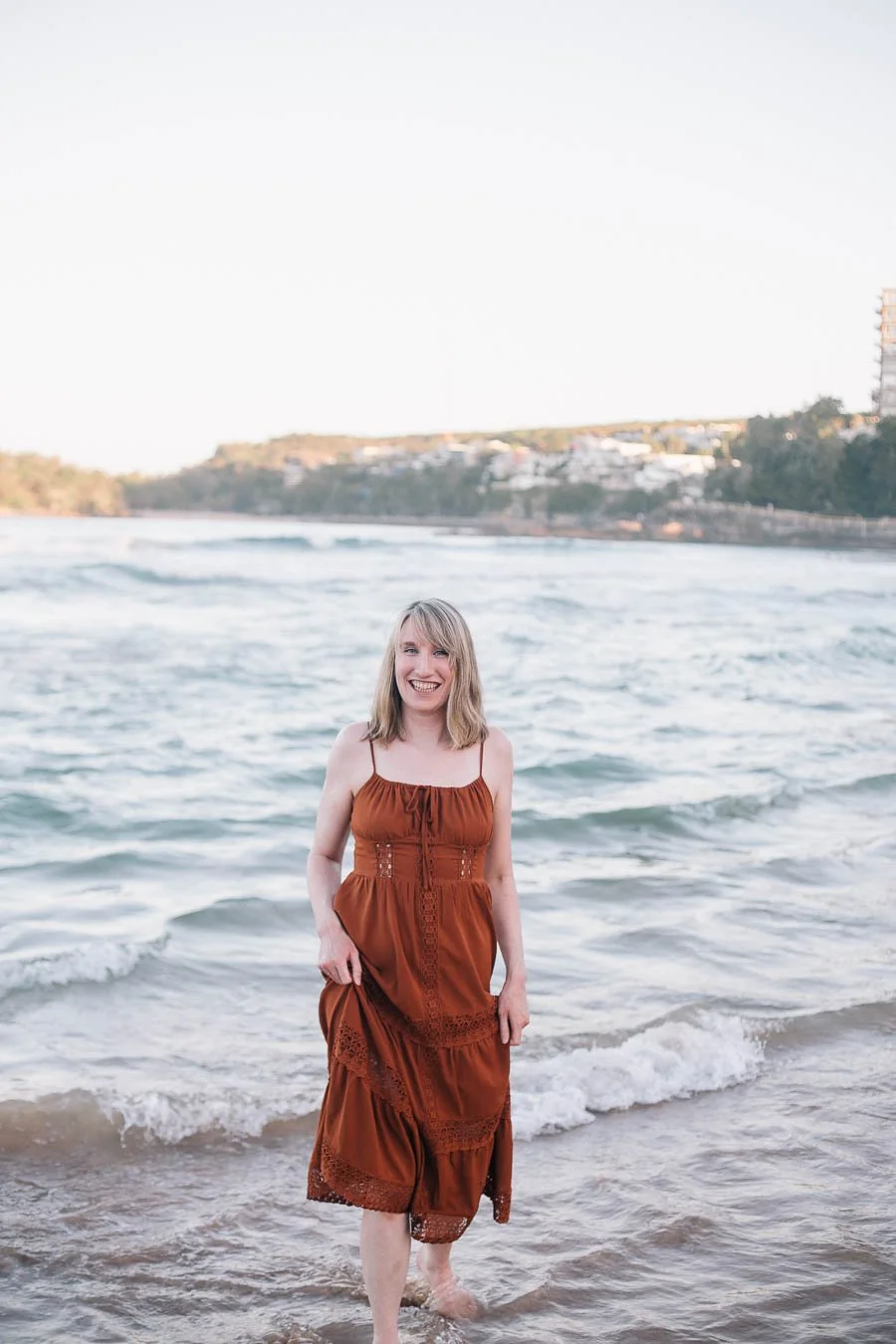 Smiling woman in a rust-colored dress standing barefoot in ocean waves at the beach
