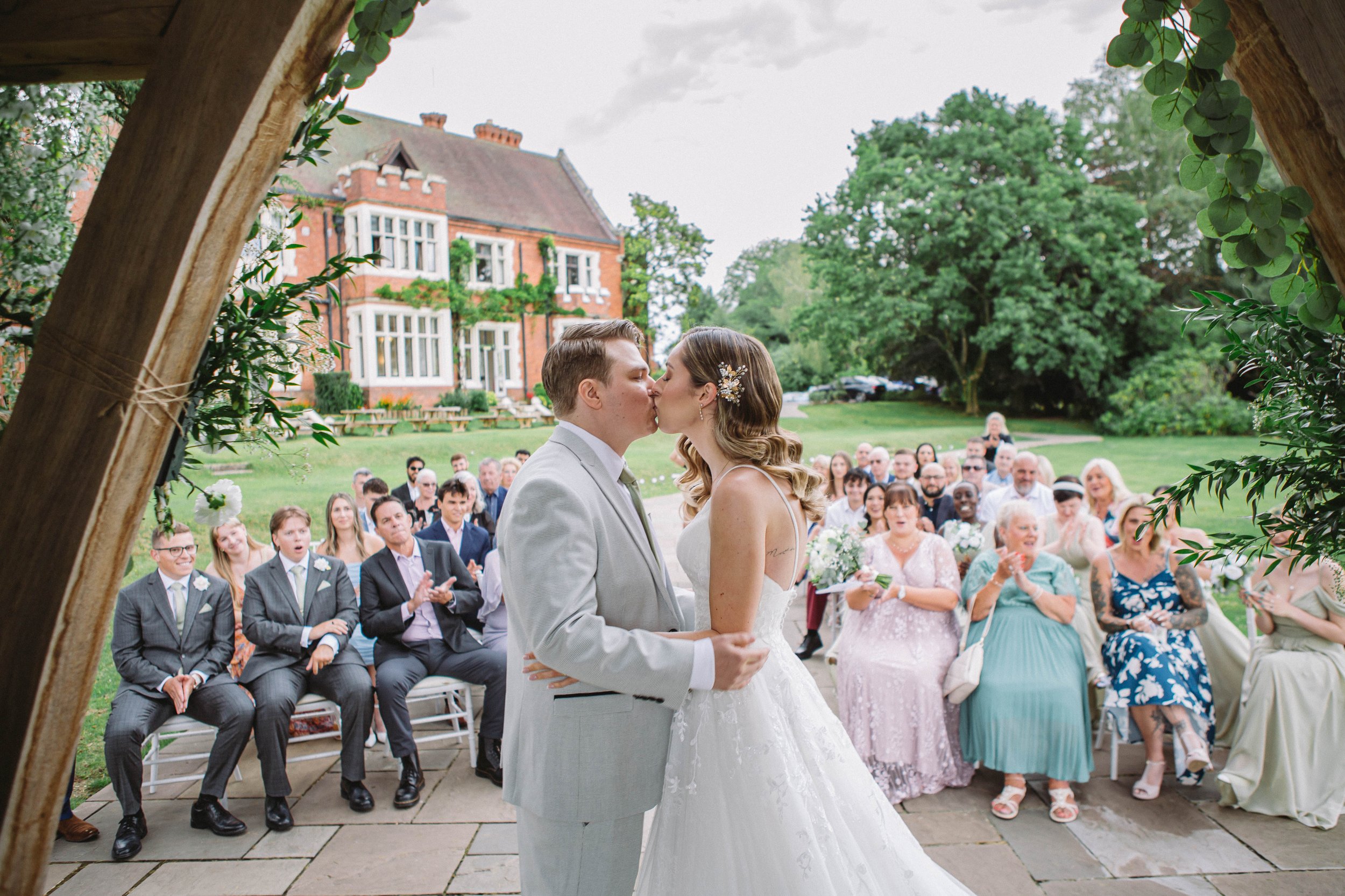 Bride and groom kiss beneath a garden arch as wedding guests applaud at an outdoor wedding ceremony.