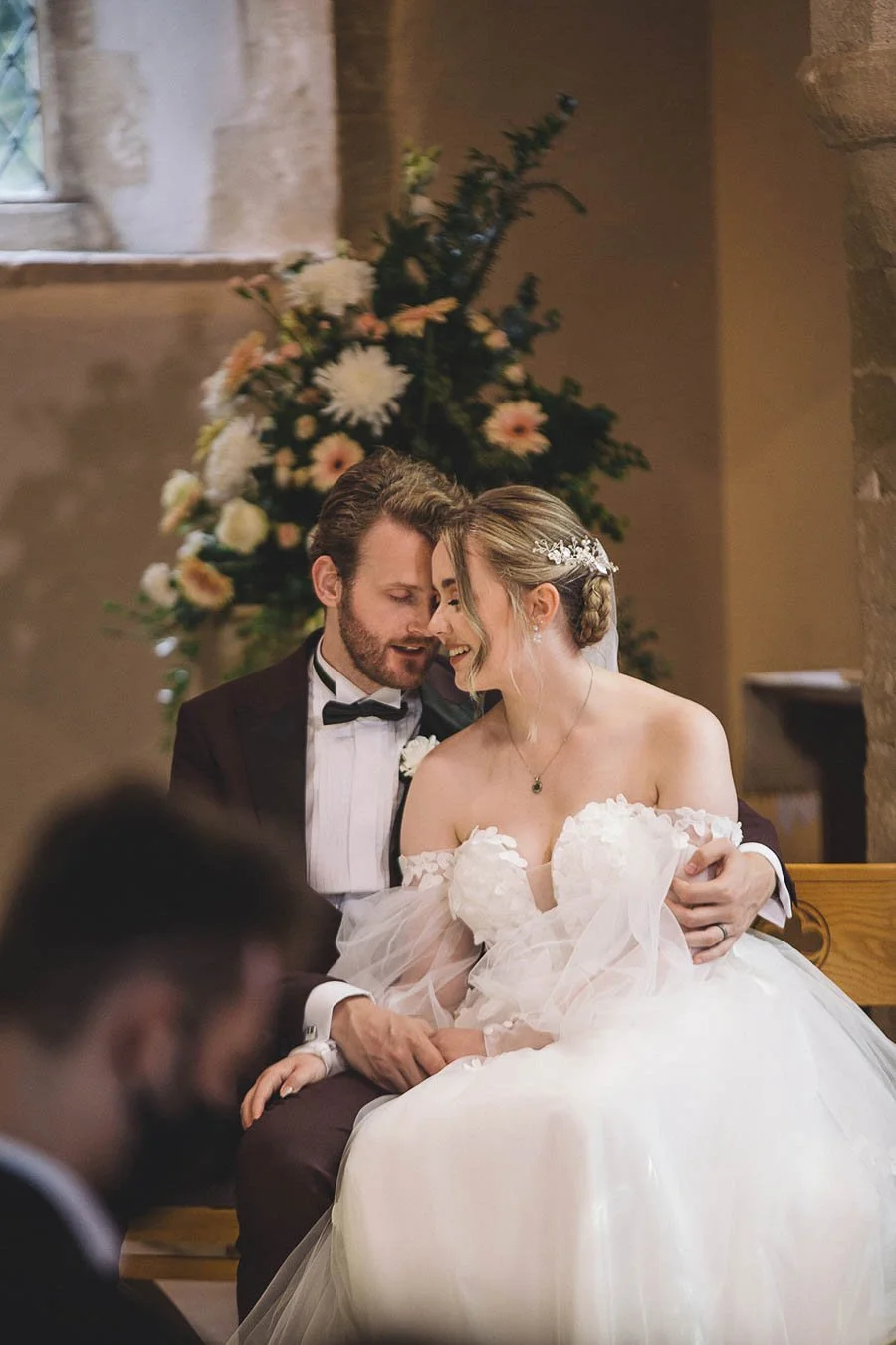 Bride and groom sitting during their wedding ceremony, leaning towards one another eyes closed. A bouquet of flowers behind them. 
